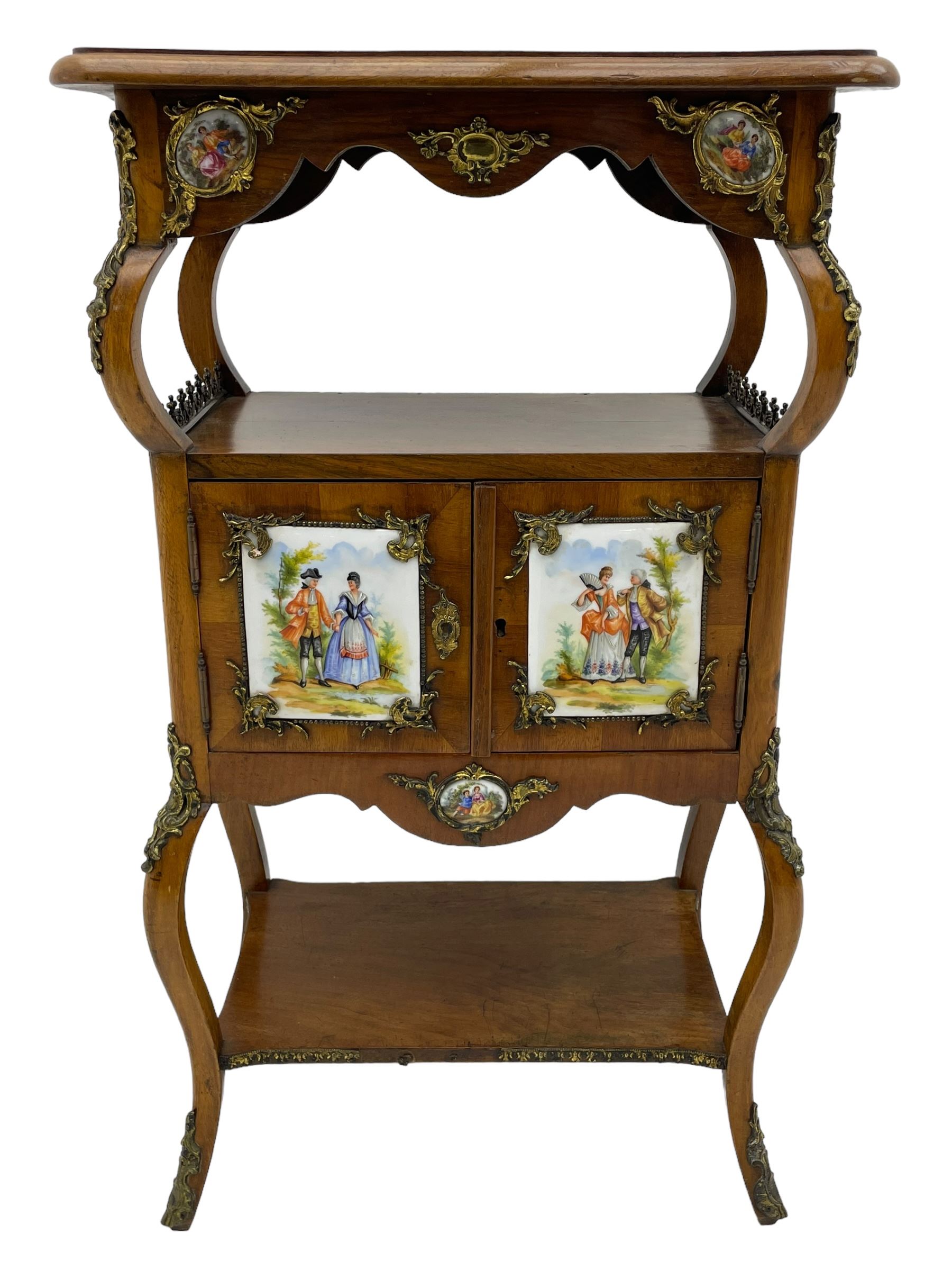 Mid-to-late 20th century French figured walnut three-tier bedside stand, shaped moulded top inlaid with figured book-matched veneers and crossbanding, double cupboard below enclosed by two doors mounted by painted porcelain panels depicting courting scenes within landscapes, on cabriole supports united by undertier, decorated with ornate cast metal cartouche mounts 