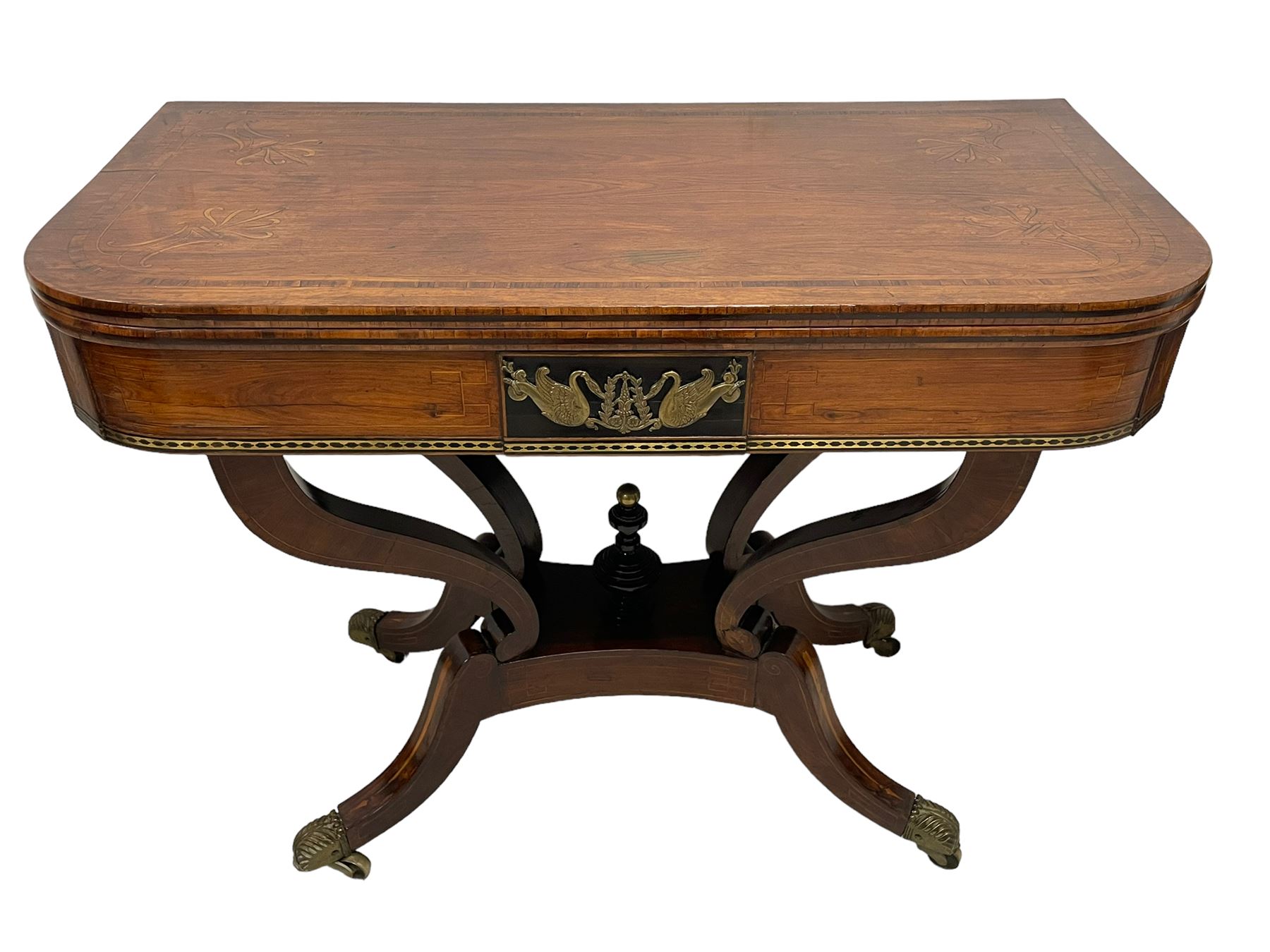 Regency inlaid rosewood card table, rectangular swivel and fold-over top with rounded corners and baize lined interior, the frieze decorated with a central ebony panel with applied gilt metal swan motifs, over a brass inlay band, raised on S-scroll supports united by concave platform base, terminating to out-splayed supports with brass cups and castors