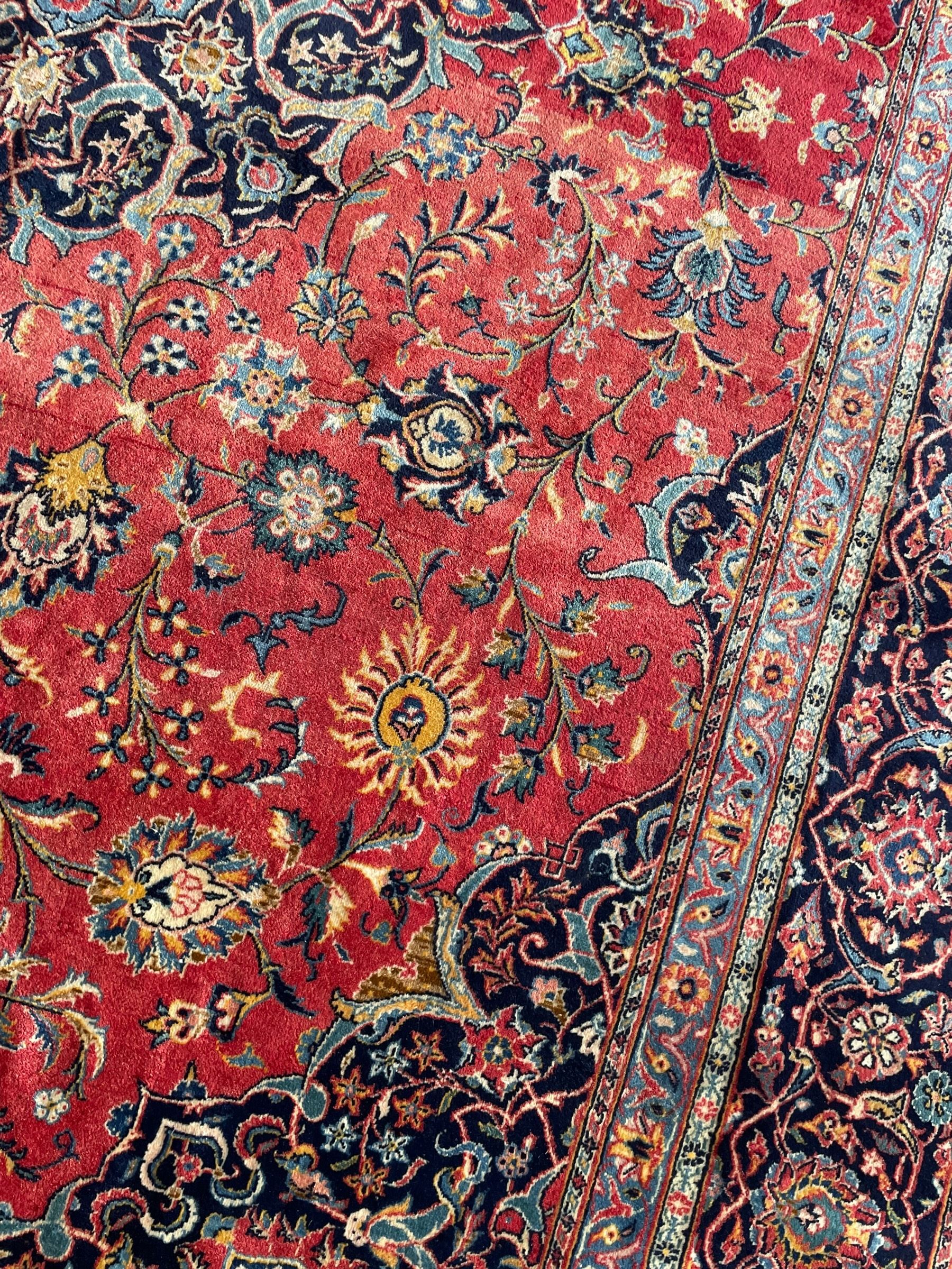 Persian Kashan crimson ground carpet, central rosette medallion on a field of trailing foliate branches decorated with palmettes, scrolling border with repeating plant motifs and flower heads, enclosed within floral pattern guard stripes