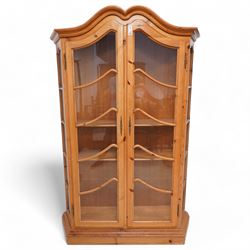 Polished pine display cabinet, shaped top over two glazed doors, fitted with three shelves, on moulded plinth base 