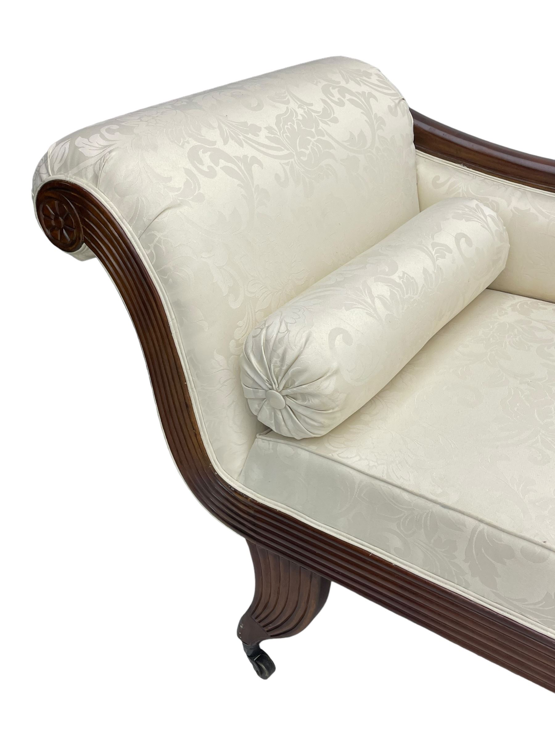 Regency design mahogany chaise longue, scrolled arms with carved rosette details, upholstered in cream damask fabric with bolster cushion, reeded frame supported by turned legs on brass castors