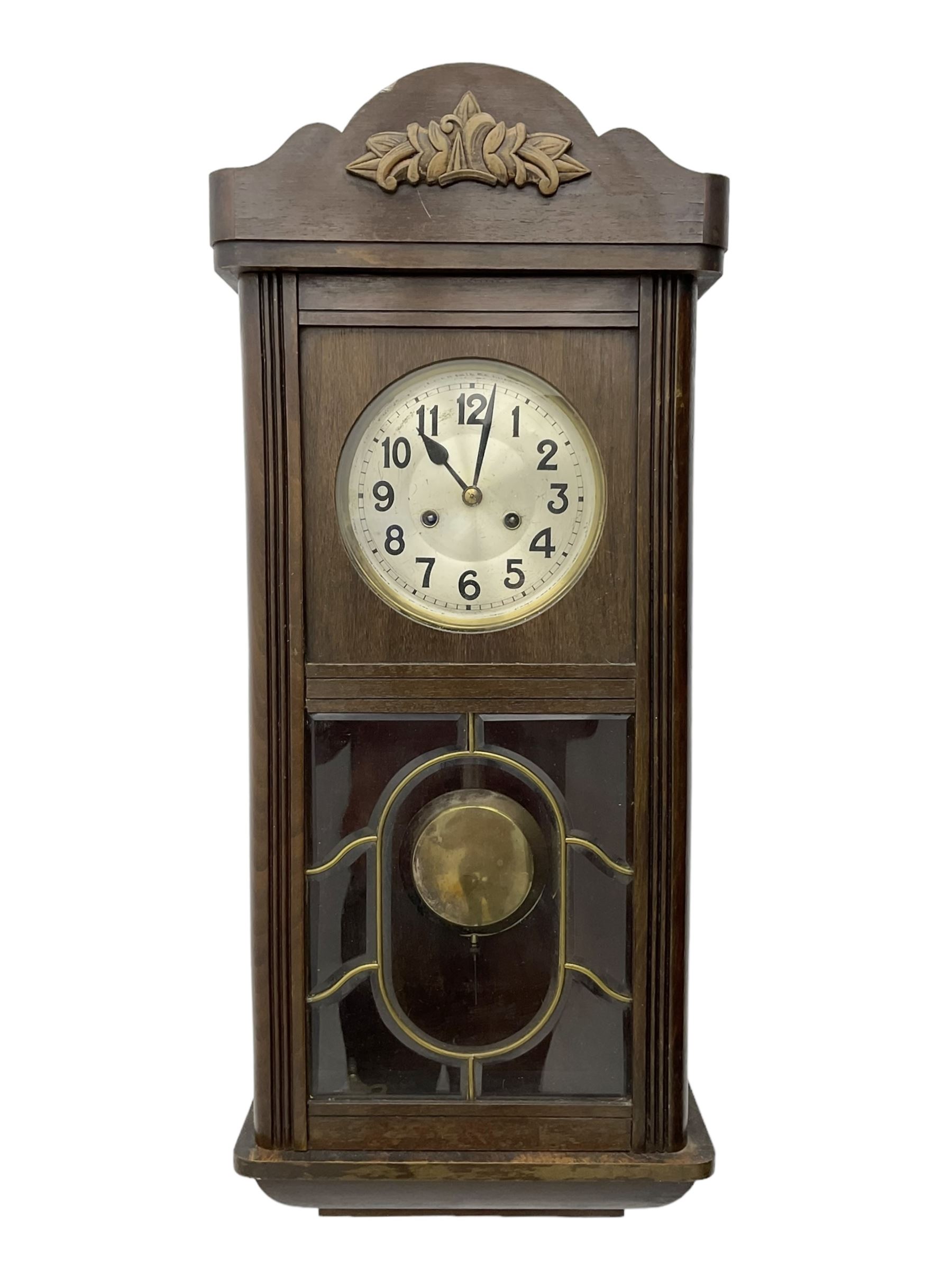 Oak cased 1930s 8-day German wall clock - with a crested top, glazed door and visible pendulum, silvered dial with Arabic numerals and spade hands, two train movement striking the hours on a gong. With pendulum and key.