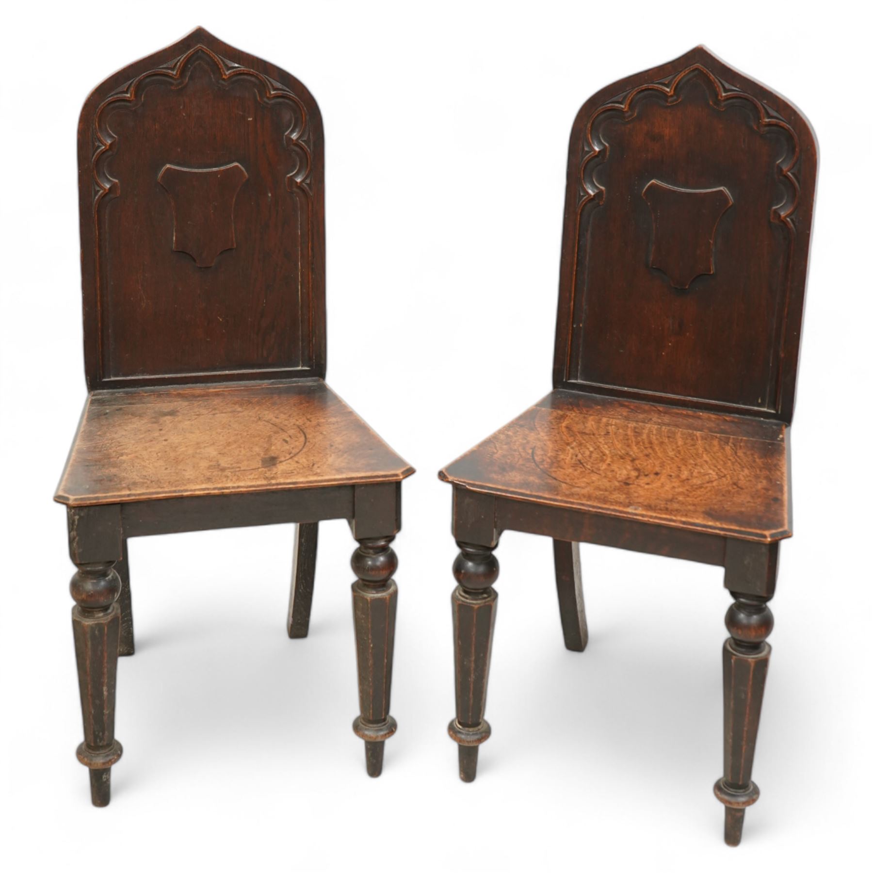 Pair of 19th century carved oak hall chairs, the arched back carved with moulded scalloped decoration with a central applied shield motif, canted panelled seat over turned octagonal supports