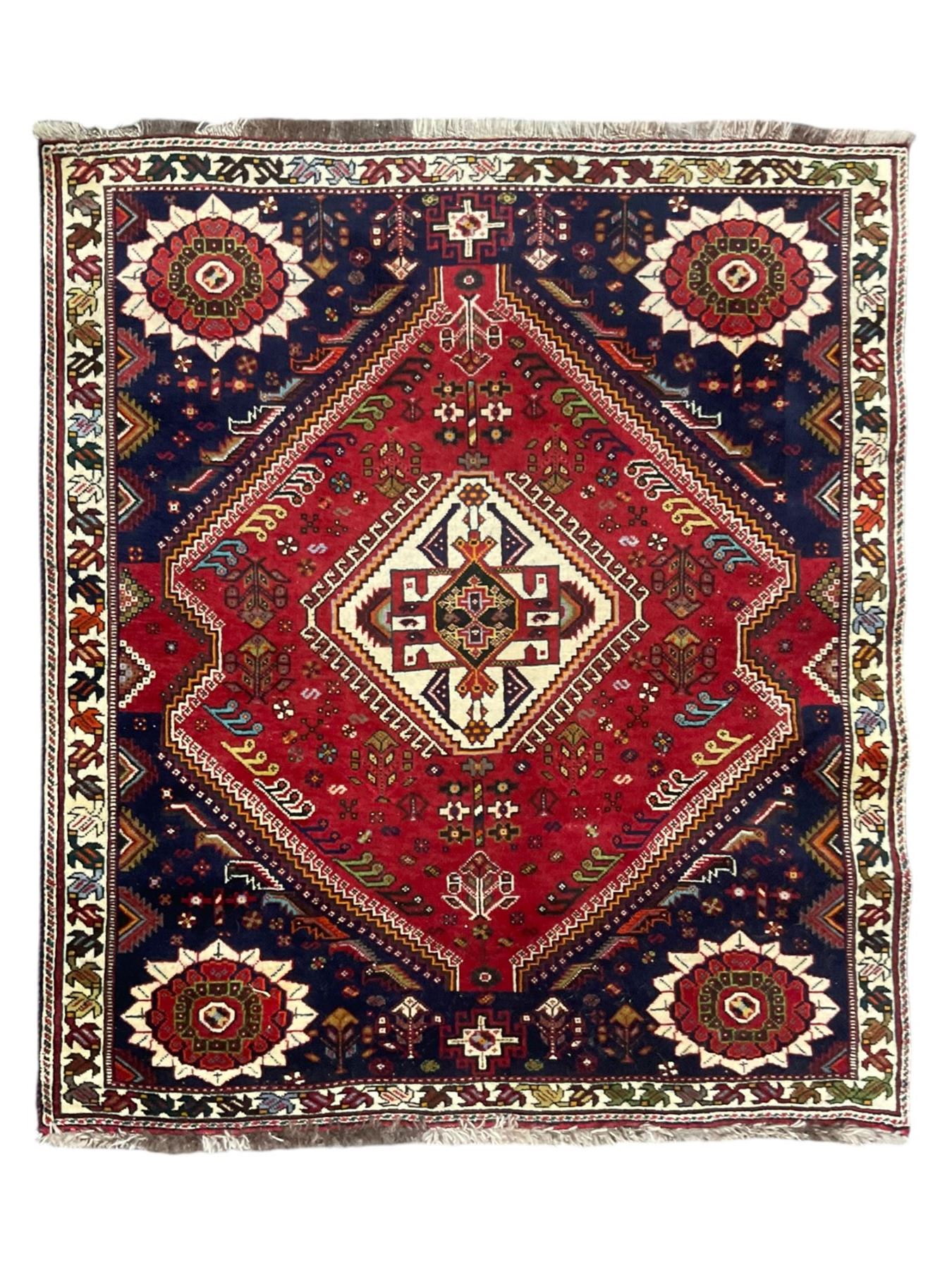 Persian Quashqai crimson ground rug, central geometric lozenge medallion surrounded by stylised plant motifs, indigo spandrels with further geometric designs, enclosed by an ivory border