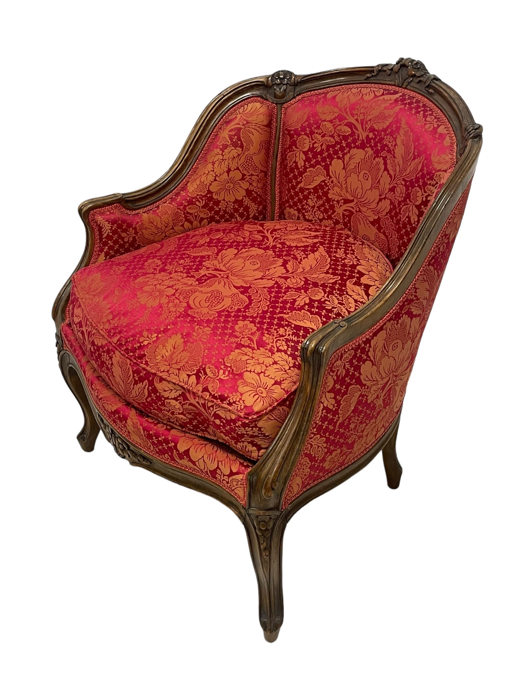 Late 20th century French design stained beech framed armchair and matching stool - the armchair with shaped and moulded frame carved with flower heads and foliage, upholstered in silk red and gold ground fabric with floral pattern, on scroll carved cabriole feet decorated with flower head carvings (W73cm, H82cm, D75cm); the footstool of concaved rectangular form with moulded frame carved with flower heads, upholstered loose cushion, on cabriole feet (70cm x 64cm, H48cm)