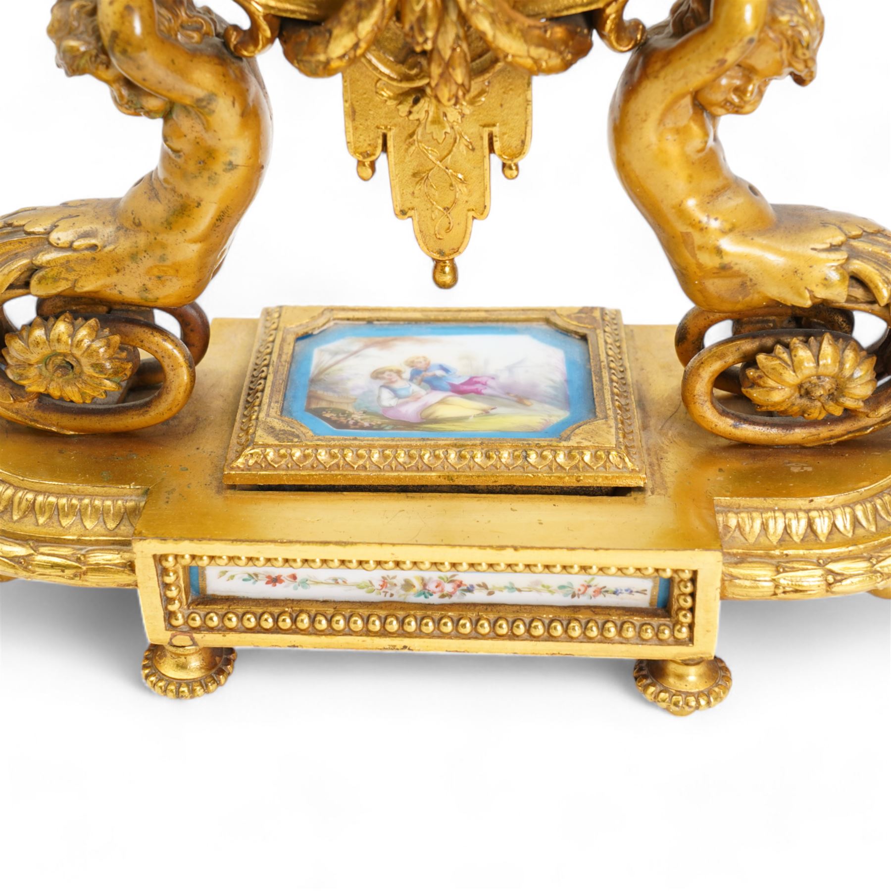 French-mid 19th century 8-day gilt and porcelain mantle clock with a break front plinth raised on paw feet, with recessed rectangular porcelain panels depicting two lovers and floral decoration, drum cased movement with a porcelain dial supported on two matching scroll supports, surmounted with a tapered porcelain urn and gilt finial, dial with cartouche Roman numerals on a blue ground and a depiction of cupid to the dial centre, twin train rack striking movement, striking the hours and half hours on a bell. With Pendulum and key.