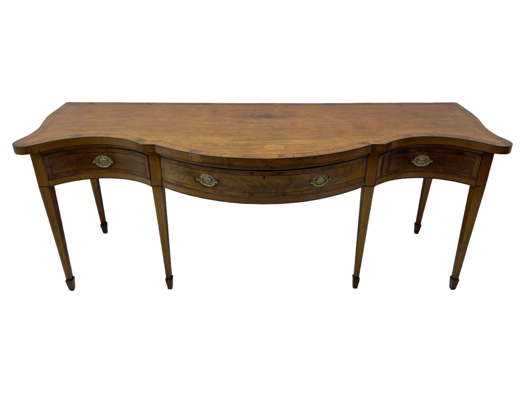 Large George III mahogany serpentine serving table, shaped top with crossing banding and stringing, the frieze fitted with three cock-beaded drawers, oval pressed brass handle plates decorated with urns, on square tapering supports with spade feet 