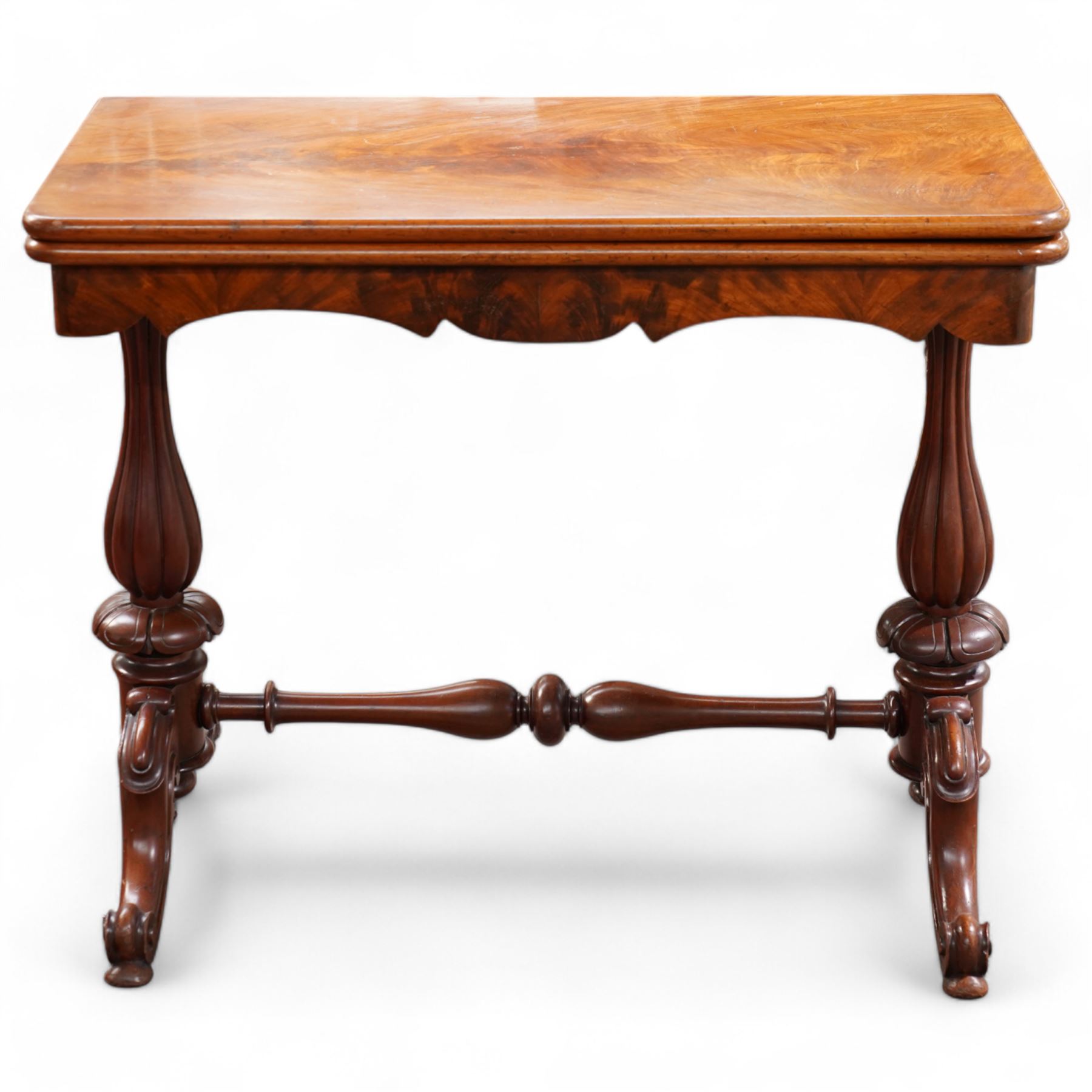 Victorian figured mahogany card table, rectangular swivel and fold-over top with rounded corners and baize lined interior, shaped apron over turned and lobe carved end supports, terminating to scroll and cartouche carved splayed supports united by swell-turned stretcher