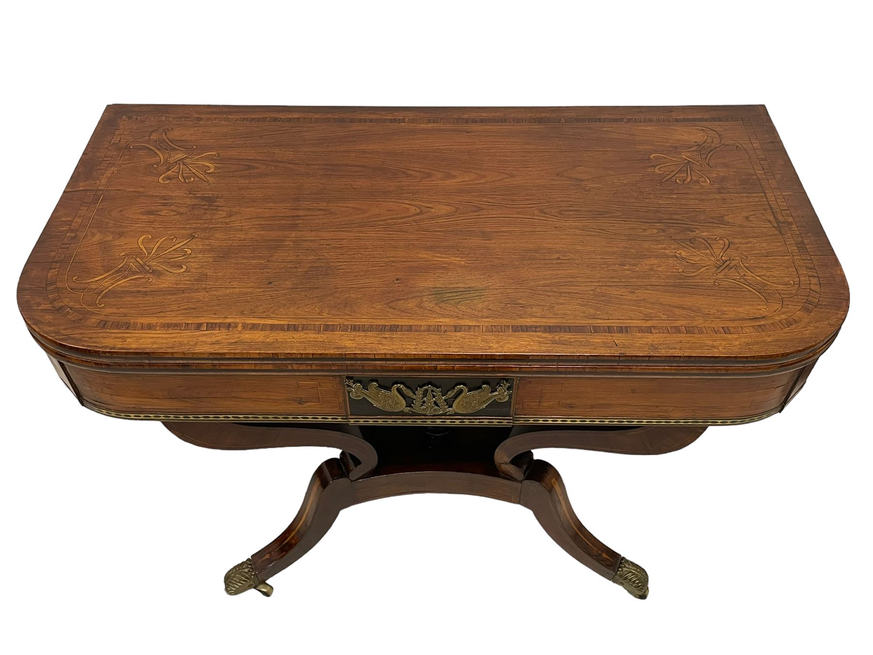 Regency inlaid rosewood card table, rectangular swivel and fold-over top with rounded corners and baize lined interior, the frieze decorated with a central ebony panel with applied gilt metal swan motifs, over a brass inlay band, raised on S-scroll supports united by concave platform base, terminating to out-splayed supports with brass cups and castors