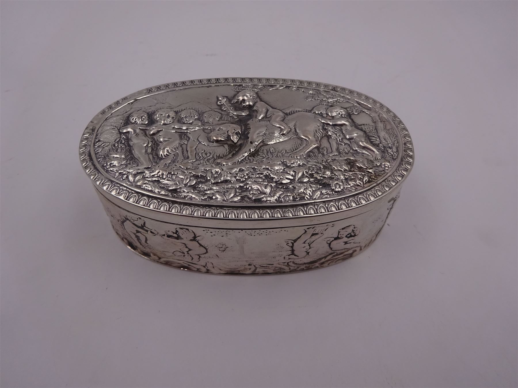 Late 19th century German Hanau silver box, of oval form, embossed throughout with putti in various scenes including riding a dog, playing instruments, pulling a cart and dancing, opening to reveal a gilt interior, with Hanau marks for B Neresheimer & Sohne, W9cm, H3cm