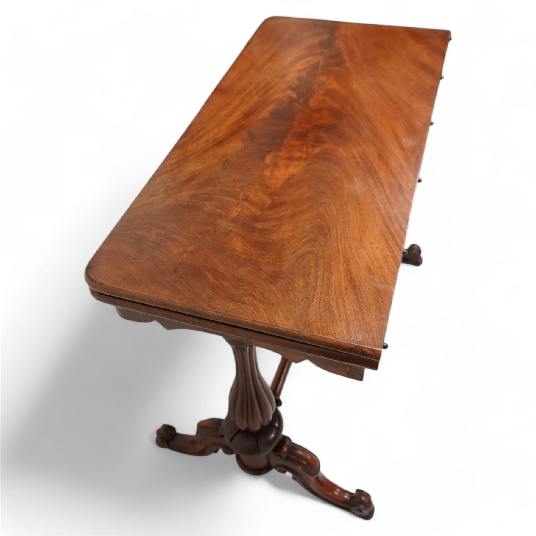 Victorian figured mahogany card table, rectangular swivel and fold-over top with rounded corners and baize lined interior, shaped apron over turned and lobe carved end supports, terminating to scroll and cartouche carved splayed supports united by swell-turned stretcher