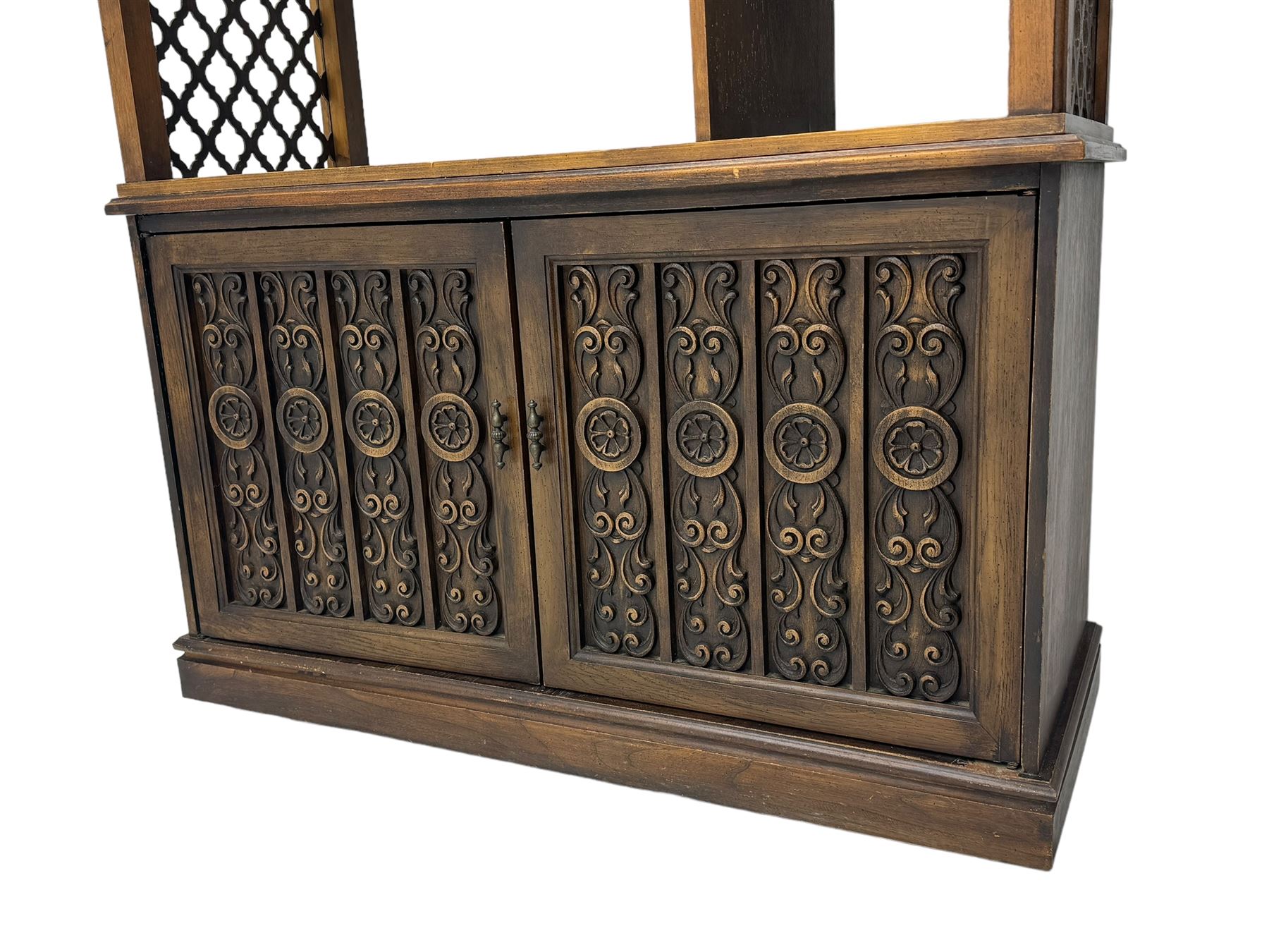 Mid 20th century oak bookcase cupboard, projecting cornice over three open shelves with subdivided sections and latticework uprights, two doors to the base with floral and scrollwork carvings and central brass handles, resting on a moulded plinth base