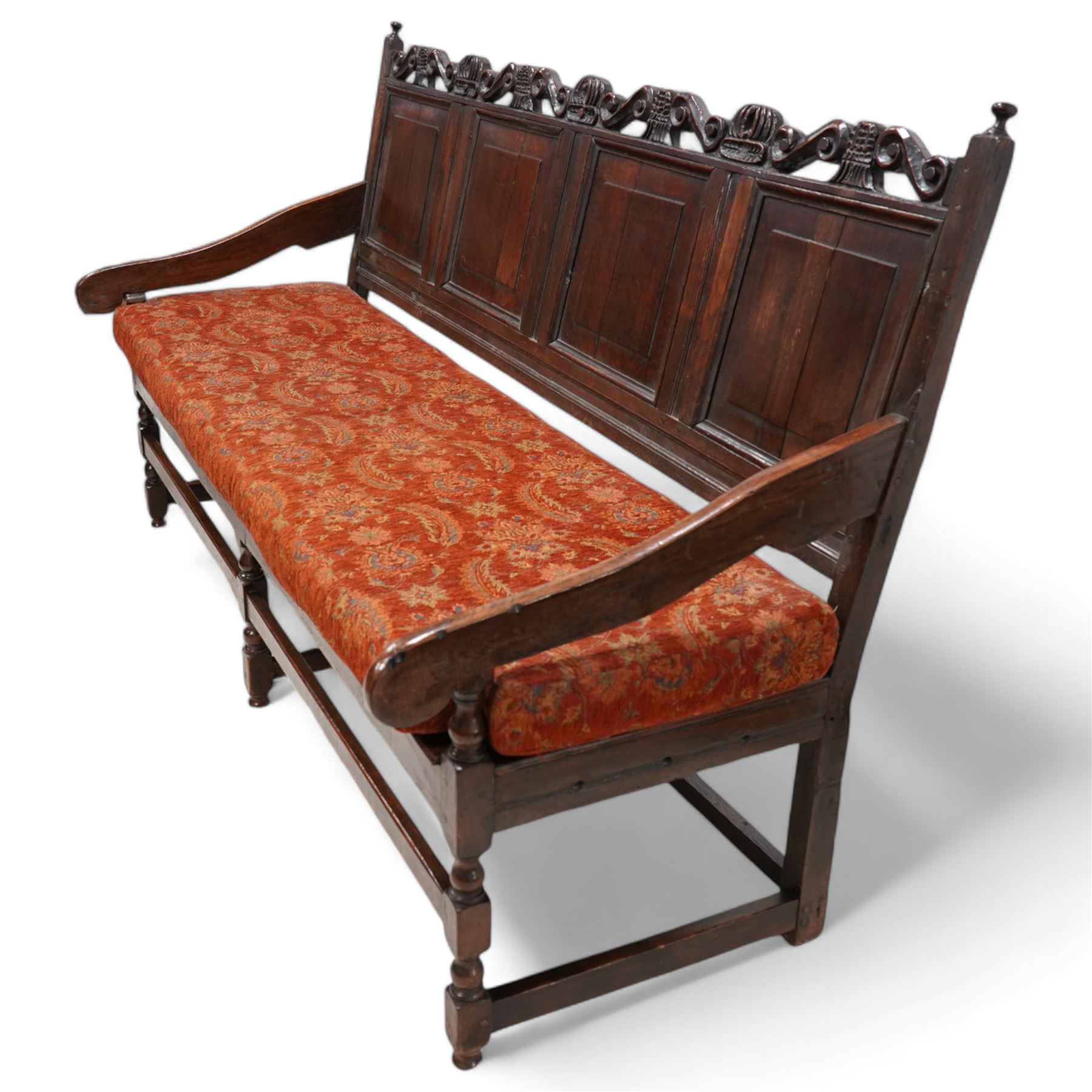 Early 18th century oak hall bench or settle, the cresting rail carved with S-scrolls and crown motifs, over a quadruple panelled back with carved rail, loose seat cushion upholstered in terracotta foliate fabric, flanked by shaped arm terminals, raised on turned supports united by stretchers