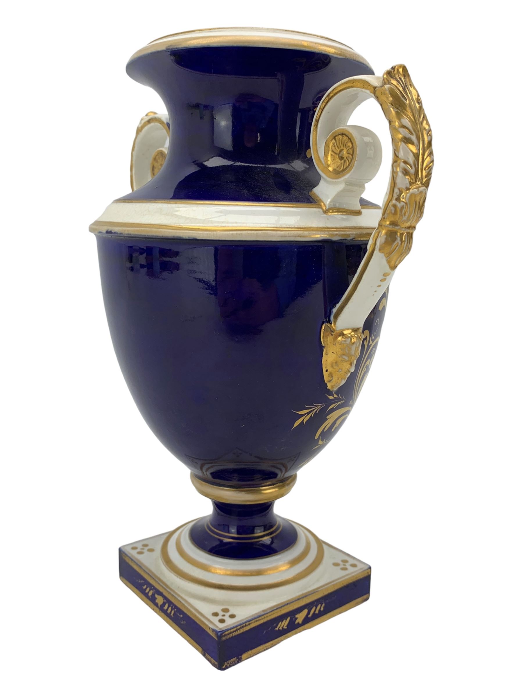 Early 19th century Derby porcelain urn, the reserve painted with a pastoral landscape and castle in the distance, with twin scroll and mask handles, upon a square pedestal base, H23cm, together with a Derby shell-shaped dish, centrally hand painted with a landscape scene titled 'In North Wales', within a cobalt blue and gilt border, L25cm (2)