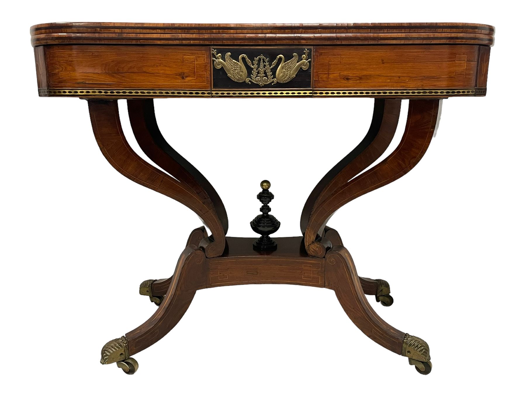 Regency inlaid rosewood card table, rectangular swivel and fold-over top with rounded corners and baize lined interior, the frieze decorated with a central ebony panel with applied gilt metal swan motifs, over a brass inlay band, raised on S-scroll supports united by concave platform base, terminating to out-splayed supports with brass cups and castors