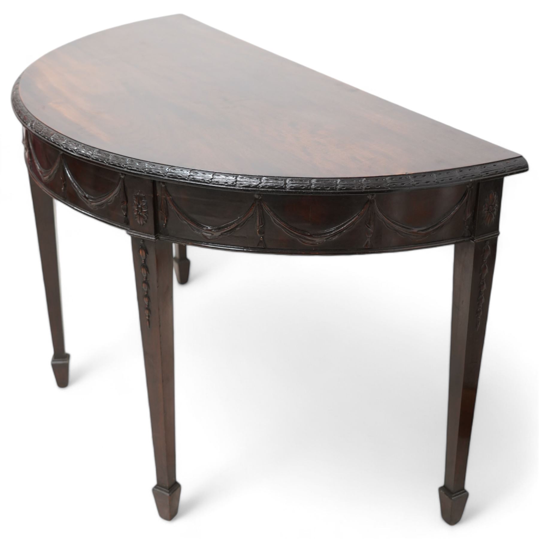 Early 20th century Hepplewhite Revival mahogany console table, demi-lune top with carved moulded edge, the frieze decorated with moulded repeating festoons, raised on square tapering supports with applied bell-flower motifs, terminating in spade feet