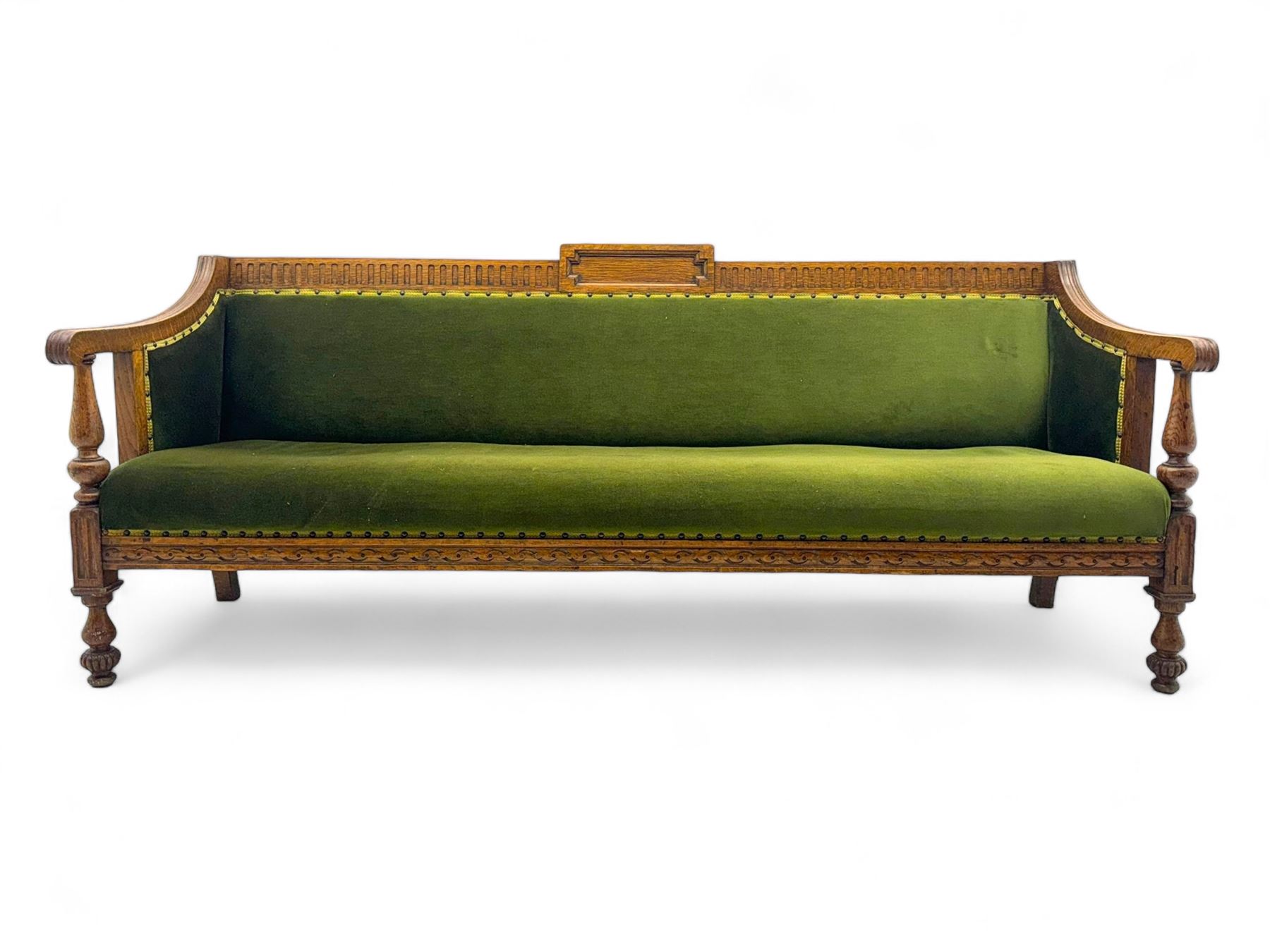 Late Victorian walnut framed sofa, rectangular backrest with carved detailing and central motif, upholstered in olive green fabric with brass studded trim and patterned front rail, scrolled fluted arms supported by turned uprights terminating in bun feet