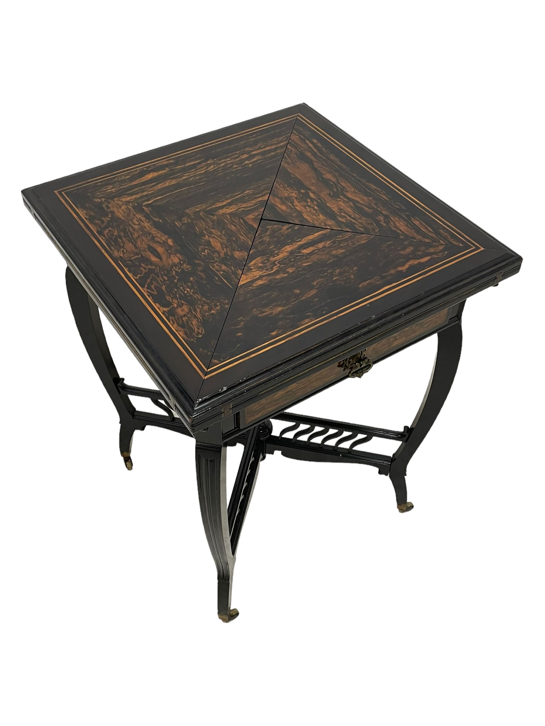 Late Victorian Coromandel wood and ebonised envelope card table, moulded swivel quadruple hinged top enclosing baize lined interior with sunken counter wells, fitted with single frieze drawer with Coromandel fascia and ornate cast handle, on four shaped and moulded supports terminating to square tapering feet, united by X-framed balustraded cross stretchers with reeded finial, on brass cups and castors 