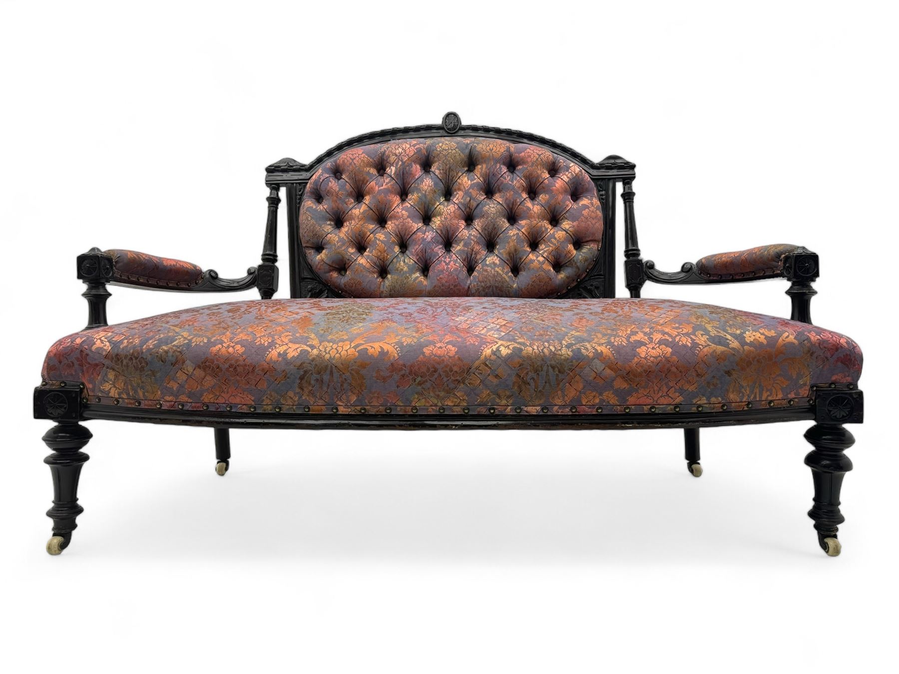 Late Victorian ebonised oak sofa, upholstered in muted purple fabric with iridescent floral pattern and nailhead trim, button-tufted backrest with carved crest rail and turned finials, scrolled padded arms supported by turned uprights, on turned supports with ceramic castors