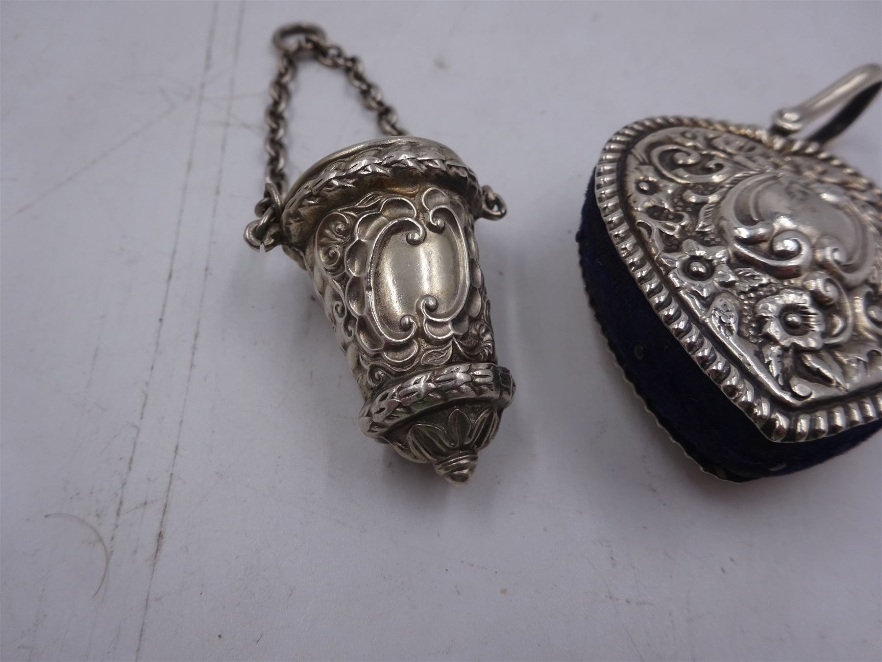 Edwardian silver pin cushion, in the form of a heart, with embossed floral decoration, with blue pin cushion between two heart shaped silver covers, with chatelain hook, hallmarked Henry Matthews, Birmingham 1902, together with a late Victorian silver thimble holder, embossed with scrolling decoration and with velvet interior, with chatelain chain, hallmarked Levi & Salaman, Birmingham 1899, pin cushion H4.5cm