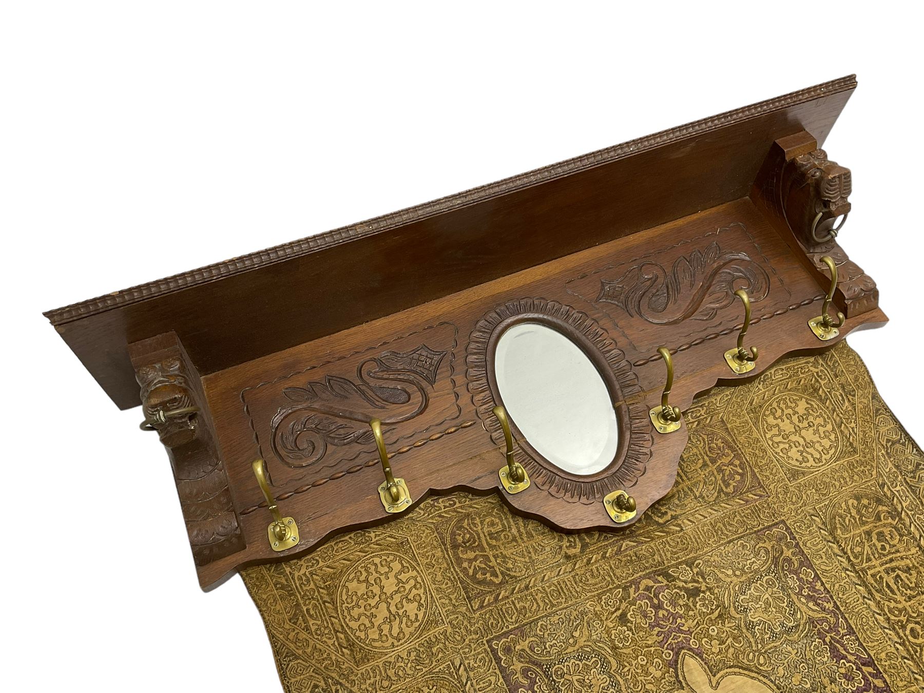 Late 20th century oak wall hanging coat rack, central bevelled oval mirror in foliate carved surround, decorated with S-scrolls and lion masks, fitted with needlework hanging 