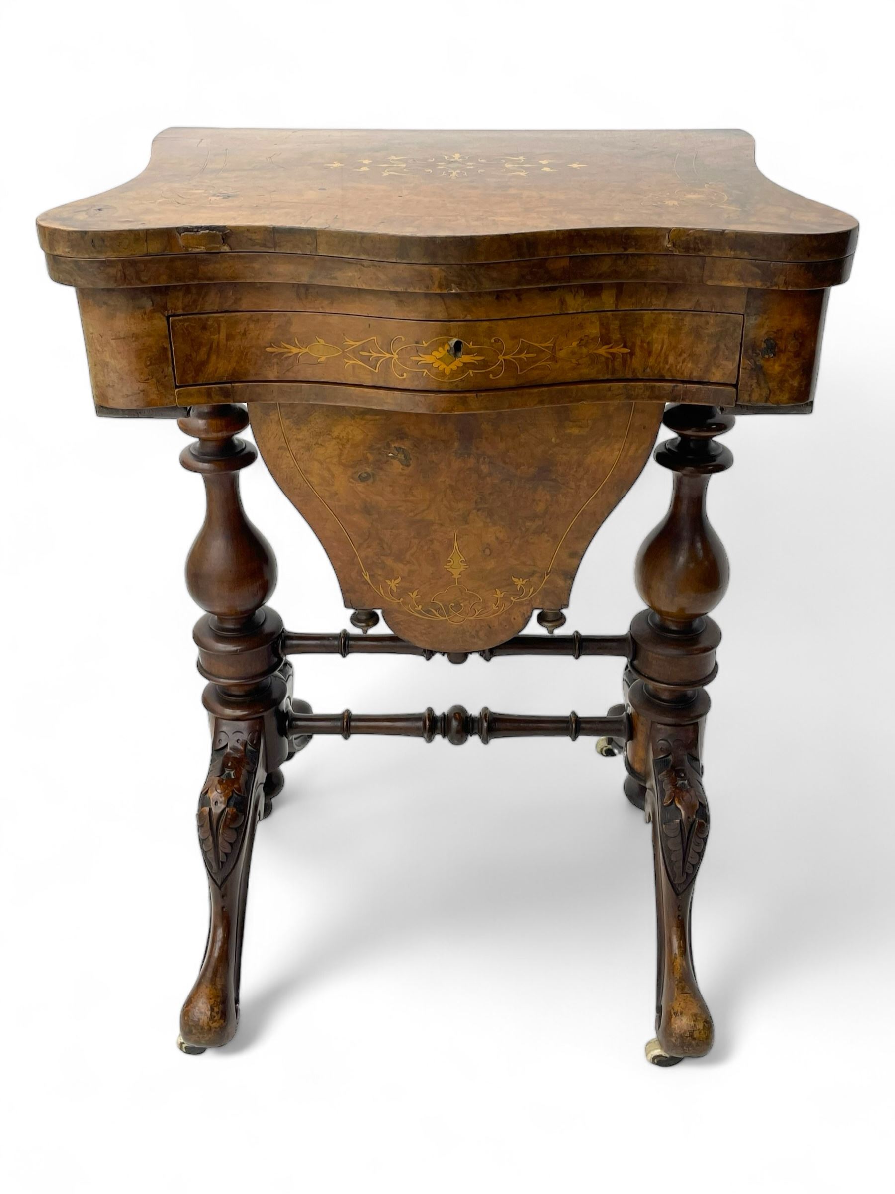 Victorian inlaid figured walnut games table, the serpentine fold-over top inlaid with scrolling foliate decoration, the top opens to reveal cribbage, backgammon and chess boards, single drawer over sliding storage well, on twin pillar supports united by double turned stretchers, on foliage carved out-splayed supports with brass and ceramic castors 