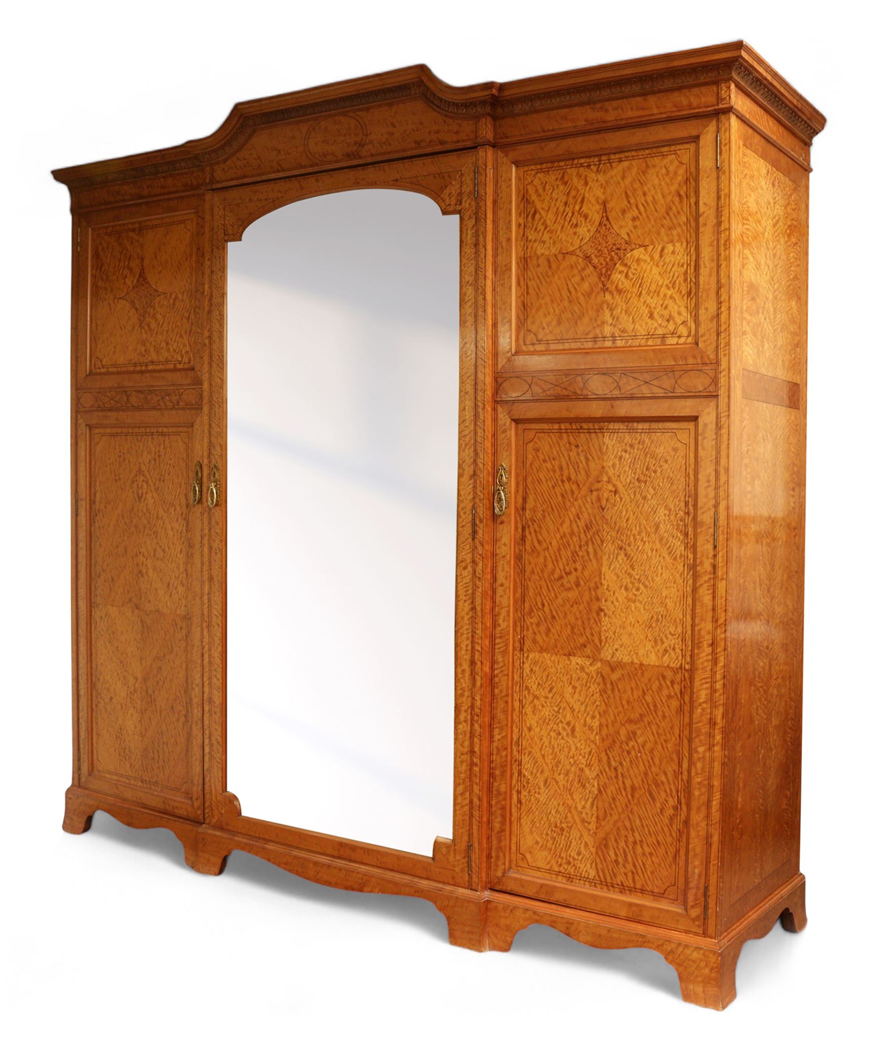 Waring & Gillow Ltd. - Edwardian inlaid satinwood triple wardrobe, stepped moulded cornice over foliate carved edge, single bevelled mirror glazed door enclosing hanging rail and three drawers, flanking panelled doors enclosing hanging rails and hooks, the door frames and panels inlaid with geometric stringing in ebony and satinwood lozenges, reeded and foliate cast loop handles and foliage cast handle plate escutcheon, quarter-matched veneers, on shaped bracket base, the locks stamped 'Gillows Lancaster' and the right hand door with metal marker's plaque 