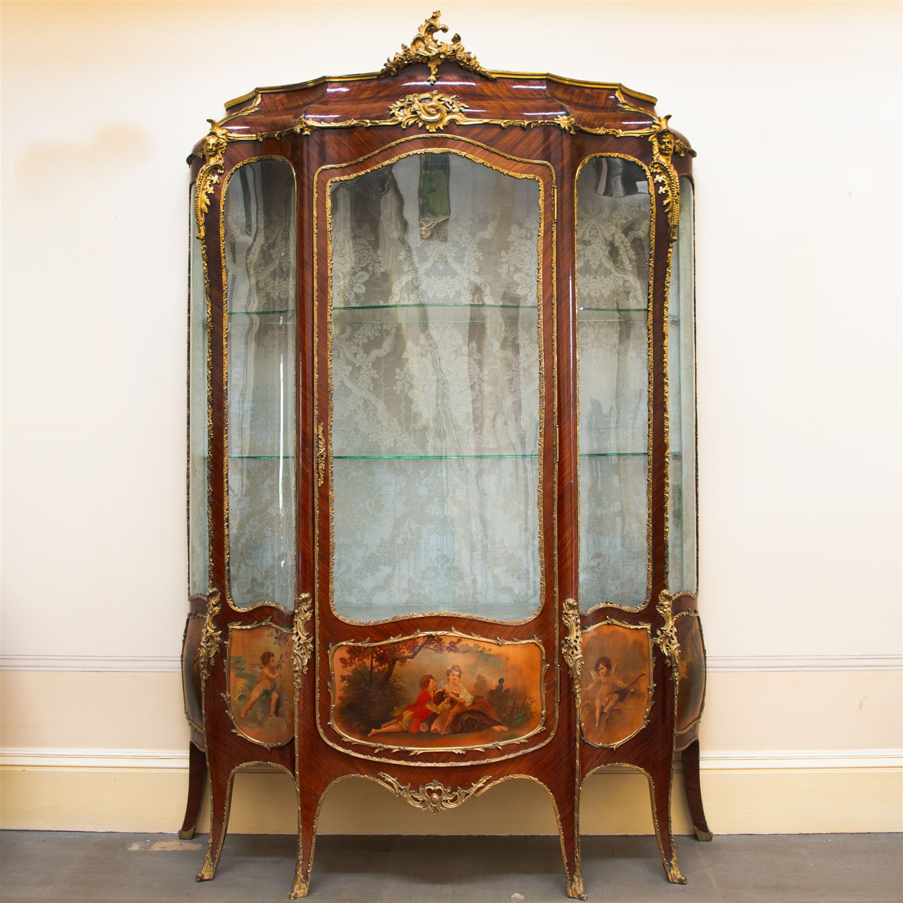 Large early 20th century French gilt metal mounted vitrine, serpentine and bombe form, C-scroll pediment decorated with extending flower heads, shaped front enclosed by single glazed door and glazed with shaped glass panes with foliate cast edging, floral pattern silk lined interior fitted with two glass shelves and one lined shelf, hand painted with Vernis Martin design panels depicting garden courting scene and putti, on cabriole supports mounted by shell and C-scroll mounts 