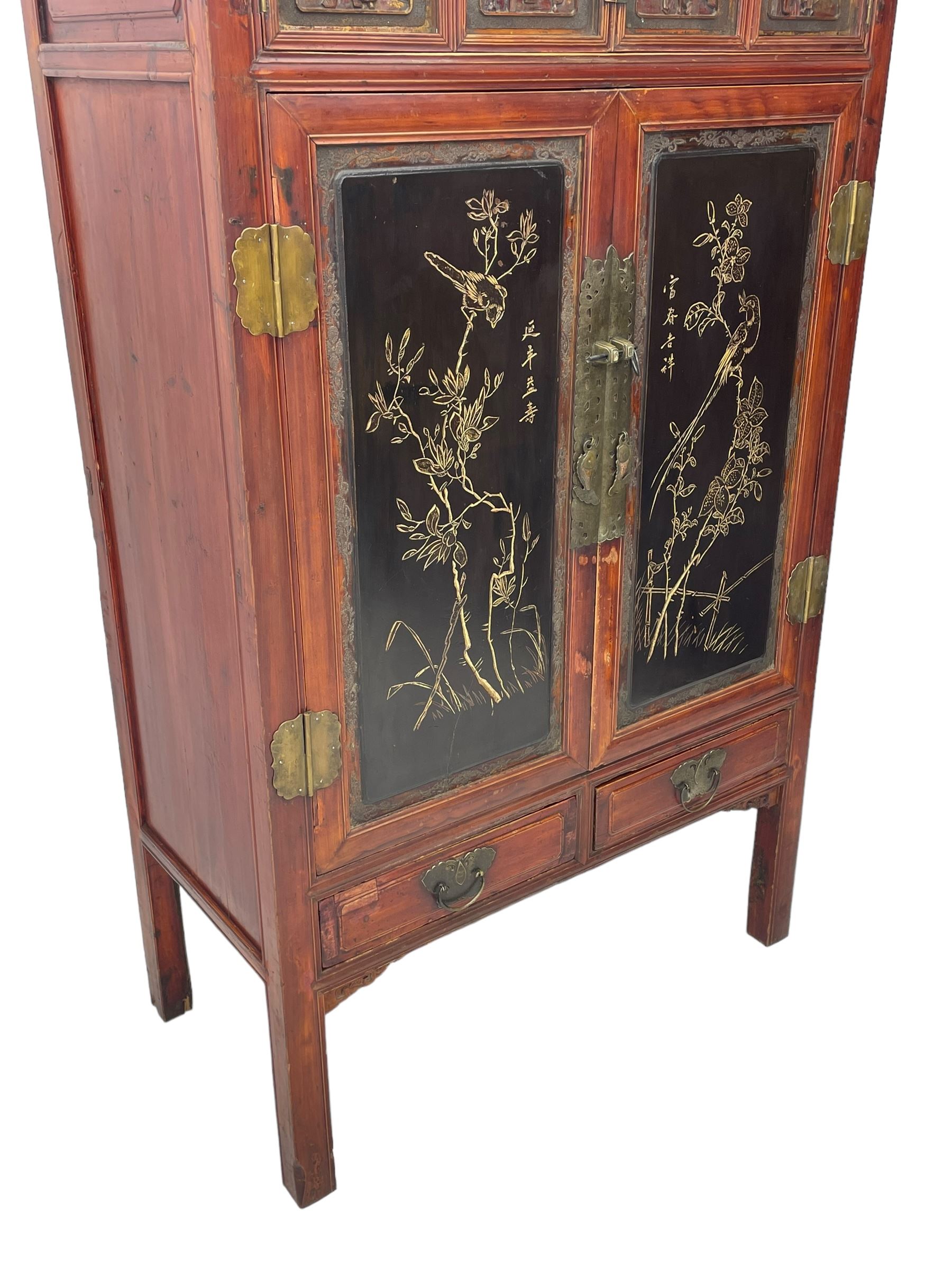 Late 19th century Chinese Qing dynasty red and black lacquered wood cabinet, Fujianese province, upper cupboard enclosed by two doors with relief carved and gilt panels depicting figures behind glass, the large cupboard enclosed by two panelled doors carved with trailing foliate branches and birds, inscribed with Chinese characters, to the left ‘Prolong life’ and to the right ‘Wealth and good fortune’, fitted with two drawers, square supports with carved brackets 