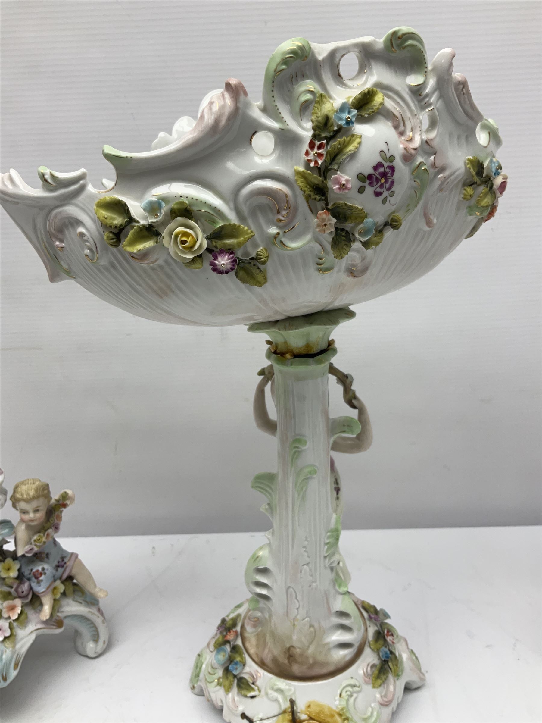 Early 20th century porcelain garniture, the central shaped basket upon a column supported by a female figure, and base with scroll feet, flanked by a pair of four branch candelabra, the scrolling branches and central stem leading to urn shaped sockets over foliate drip pans, upon shaped columns and bases with applied putto, the whole with encrusted flowers throughout, basket H33.5cm, candelabra H52cm