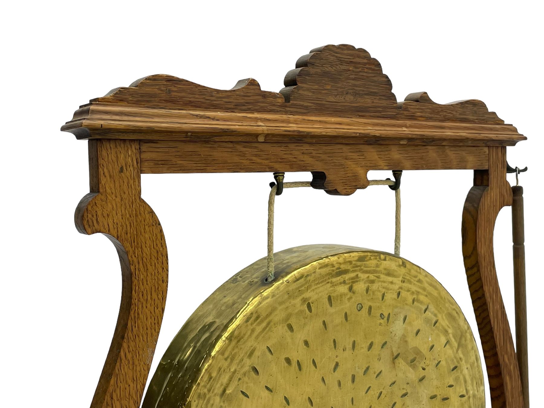 Early 20th century oak gong stand with gong and hammer, shaped cresting rail over moulded upper edge on shaped supports, hanging brass drum gong over fretwork middle rail, on raised platforms terminating to splayed feet