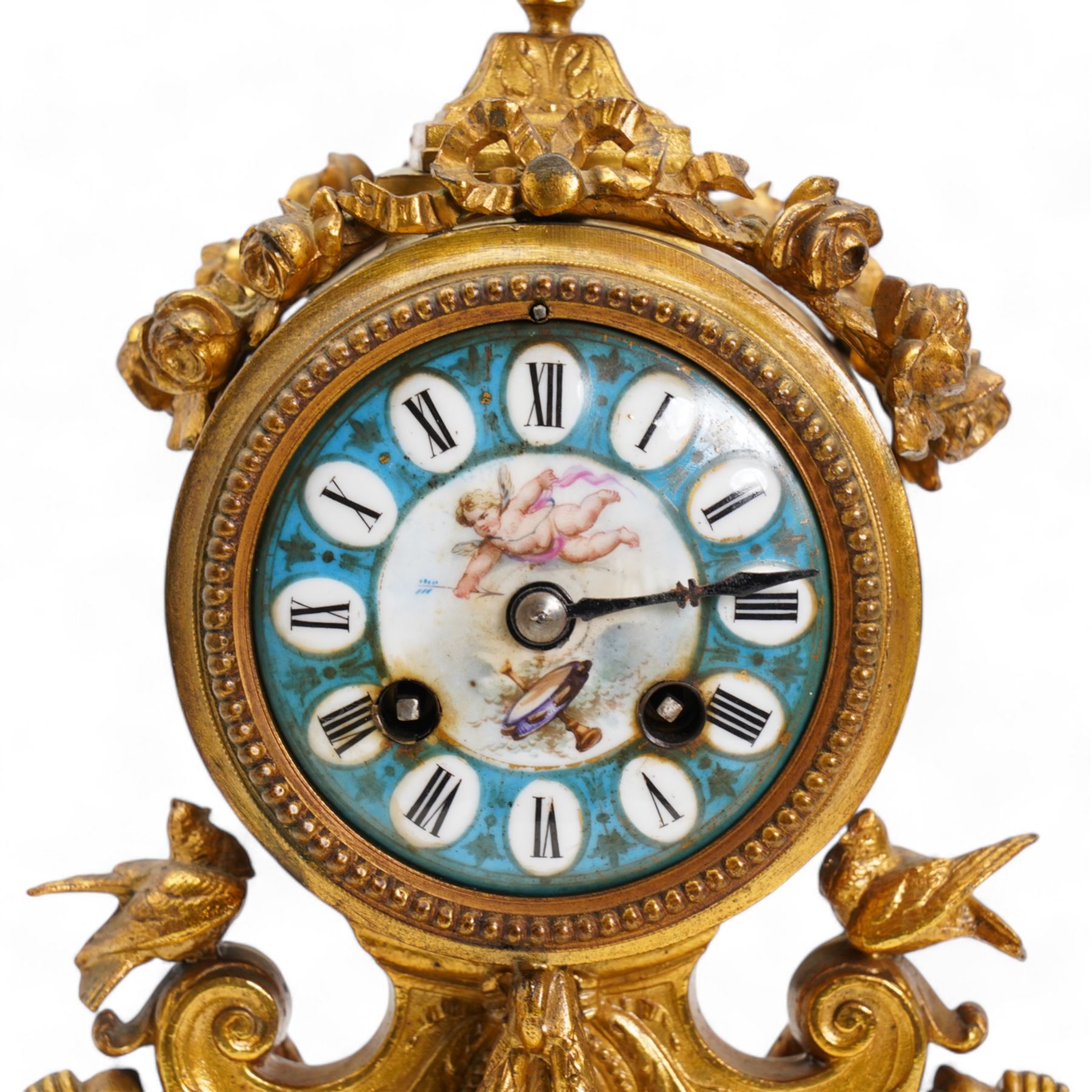 French-mid 19th century 8-day gilt and porcelain mantle clock with a break front plinth raised on paw feet, with recessed rectangular porcelain panels depicting two lovers and floral decoration, drum cased movement with a porcelain dial supported on two matching scroll supports, surmounted with a tapered porcelain urn and gilt finial, dial with cartouche Roman numerals on a blue ground and a depiction of cupid to the dial centre, twin train rack striking movement, striking the hours and half hours on a bell. With Pendulum and key.