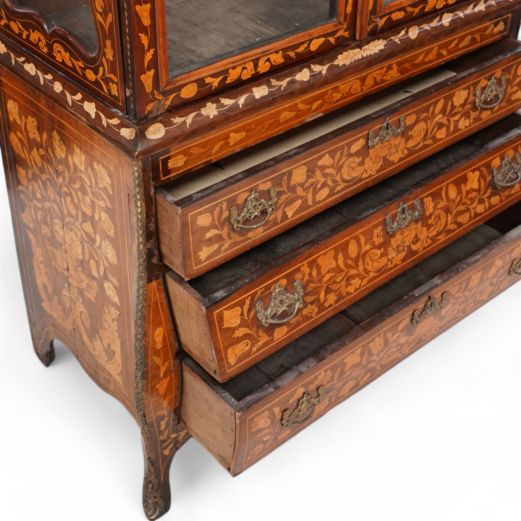 18th century Dutch marquetry walnut and gilt metal mounted vitrine cabinet on chest, shaped pediment carved with scrolling foliage cartouche, inlaid with trailing leafy branches and flower heads, two internal shelves enclosed by two glazed doors, the lower section with bombe outline fitted with three drawers, with ornate cast gilt metal edging and terminal caps