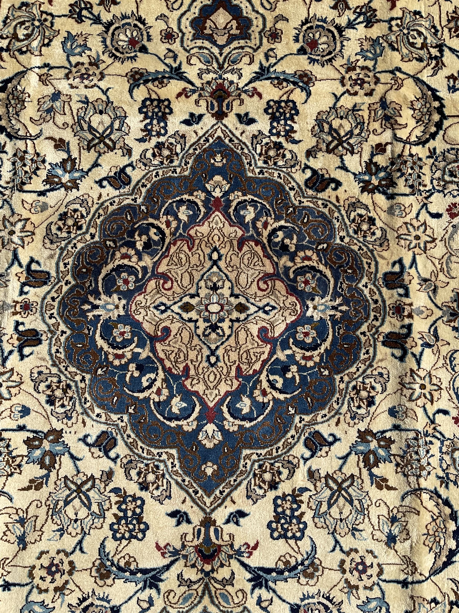 Persian Nain ivory ground carpet, central indigo medallion, the busy floral field decorated with scrolling vines and flower heads in shades of blue, beige, and red, wide guarded border with repeating palmettes and foliate motifs