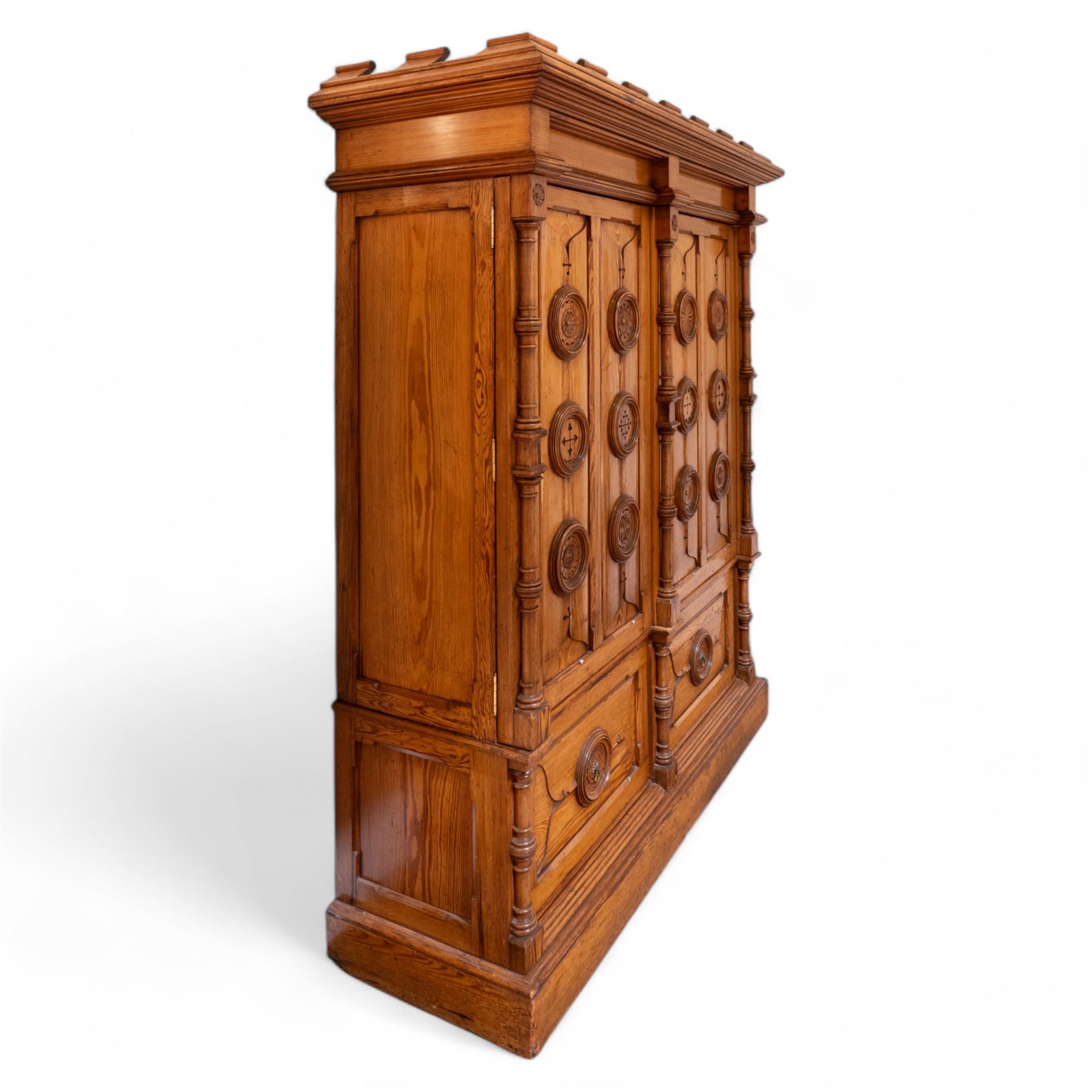 19th century pitch pine ecclesiastical vestment wardrobe or sacristy cabinet, crenelated and projecting moulded cornice over two panelled doors, the panelled doors carved with linen folds and mounted with rows of roundels depicting crucifixes and flower heads, enclosed by turned pilasters, two drawers to the base, on moulded plinth base 