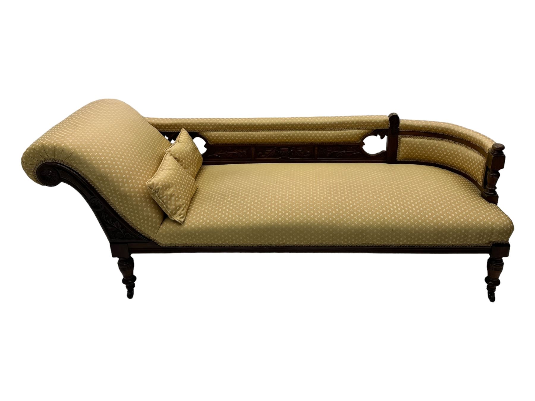 Late Victorian walnut framed chaise longue, scrolled backrest and sides upholstered in patterned yellow fabric, carved floral motifs to the backrest and sides, raised on turned supports with castors