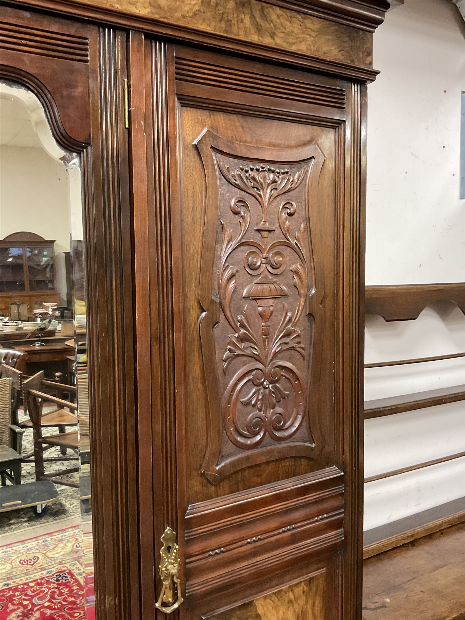 Late Victorian walnut triple wardrobe, the pierced broken swan neck pediment carved with foliate C-scrolls, central bevelled mirror door flanked by two panelled doors, the panels carved with urns, C-scrolls and trailing foliage, the interior fitted with hanging rails, long and short drawer, on moulded plinth base