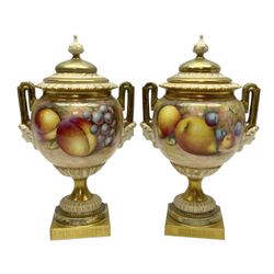 Pair of mid 20th century Royal Worcester vases and covers decorated by Alan Telford, each of ovoid form with twin mask mounted handles, and gilt covers with bud finials, upon a gilt circular pedestal foot and gilt square plinth, the body hand painted with a still life of fruit upon a mossy ground, signed Telford, with black printed mark beneath and painted shape number 2363, H21cm