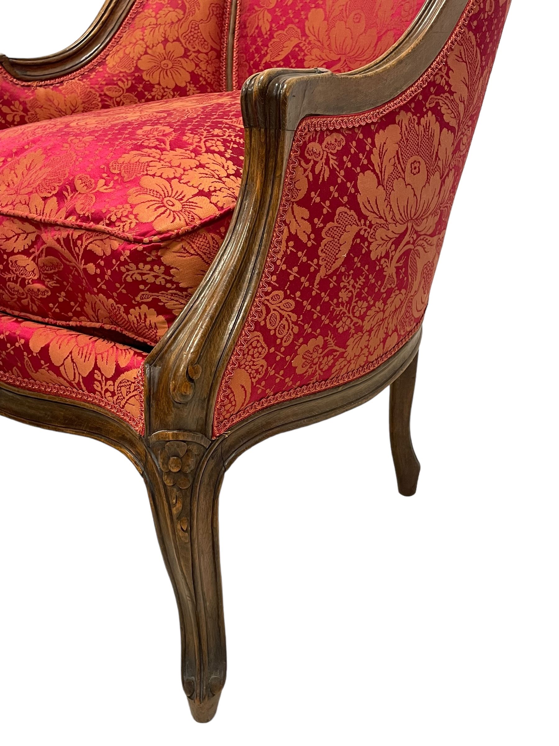 Late 20th century French design stained beech framed armchair and matching stool - the armchair with shaped and moulded frame carved with flower heads and foliage, upholstered in silk red and gold ground fabric with floral pattern, on scroll carved cabriole feet decorated with flower head carvings (W73cm, H82cm, D75cm); the footstool of concaved rectangular form with moulded frame carved with flower heads, upholstered loose cushion, on cabriole feet (70cm x 64cm, H48cm)