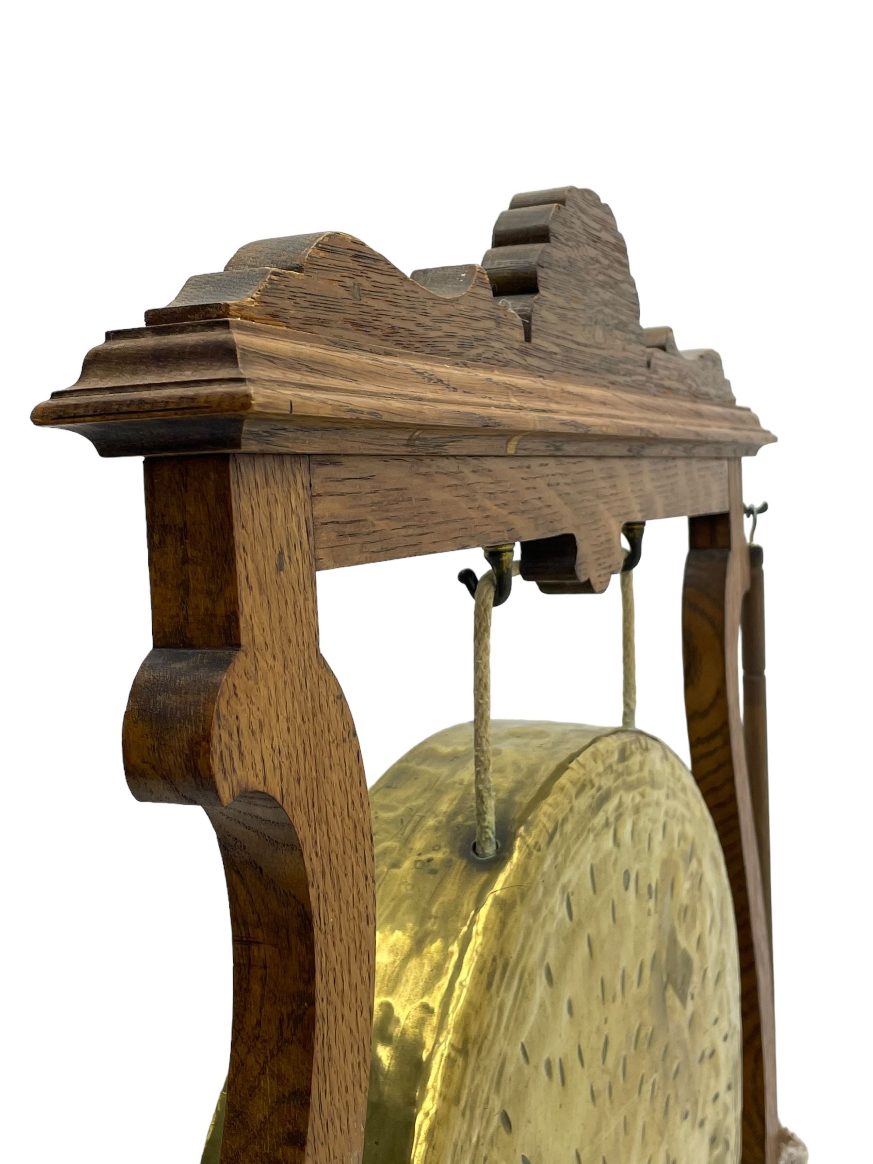 Early 20th century oak gong stand with gong and hammer, shaped cresting rail over moulded upper edge on shaped supports, hanging brass drum gong over fretwork middle rail, on raised platforms terminating to splayed feet