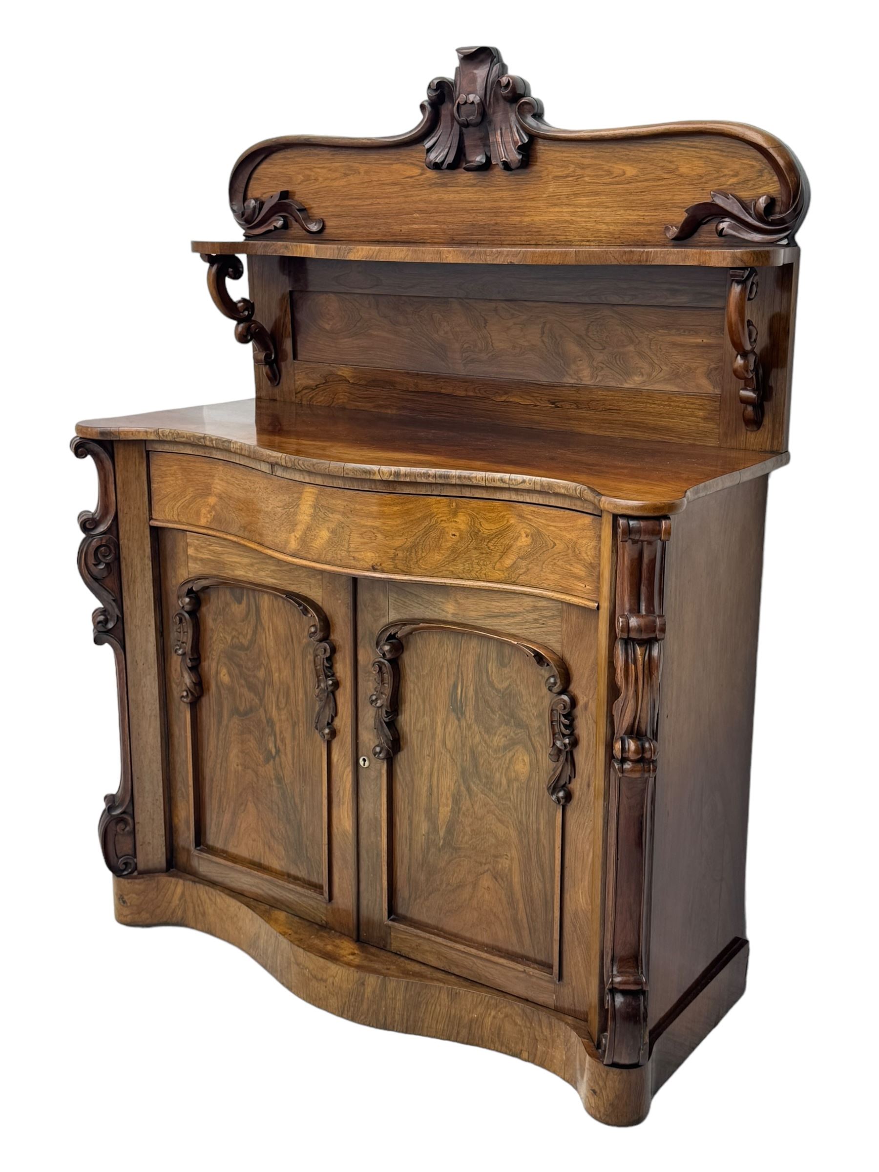 Mid-19th century rosewood chiffonier, raised back with scroll and leaf carved shaped pediment, serpentine top over drawer and double cupboard with scrollwork decoration, on shaped plinth base