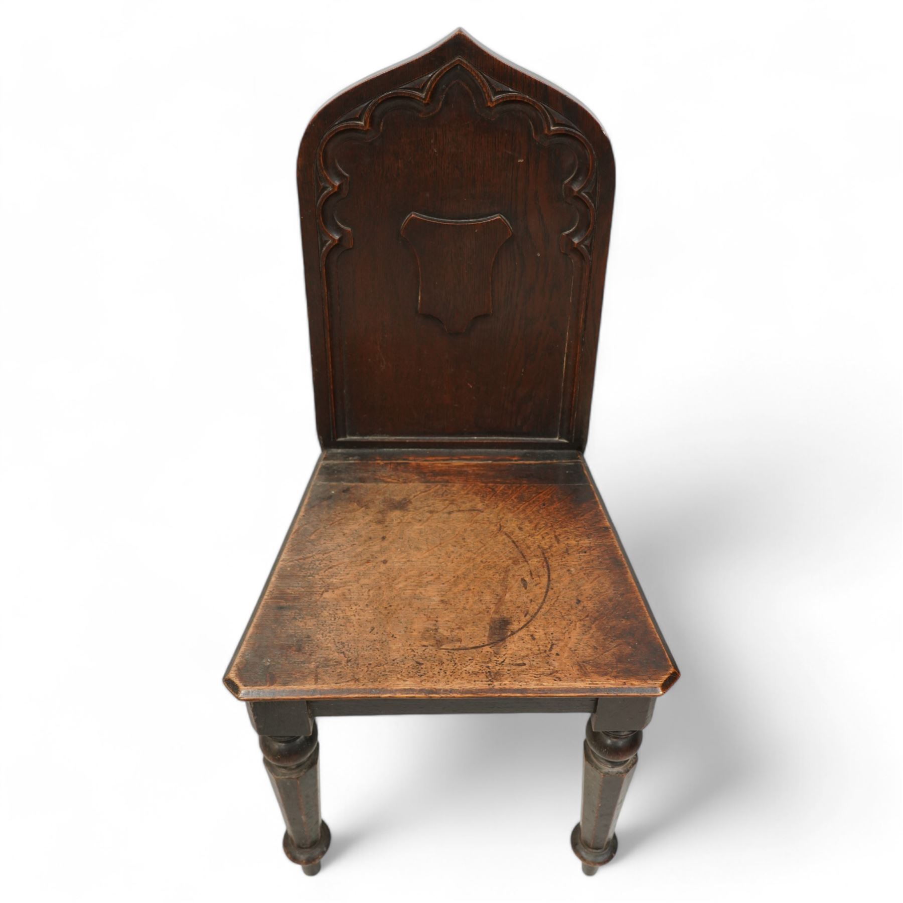 Pair of 19th century carved oak hall chairs, the arched back carved with moulded scalloped decoration with a central applied shield motif, canted panelled seat over turned octagonal supports