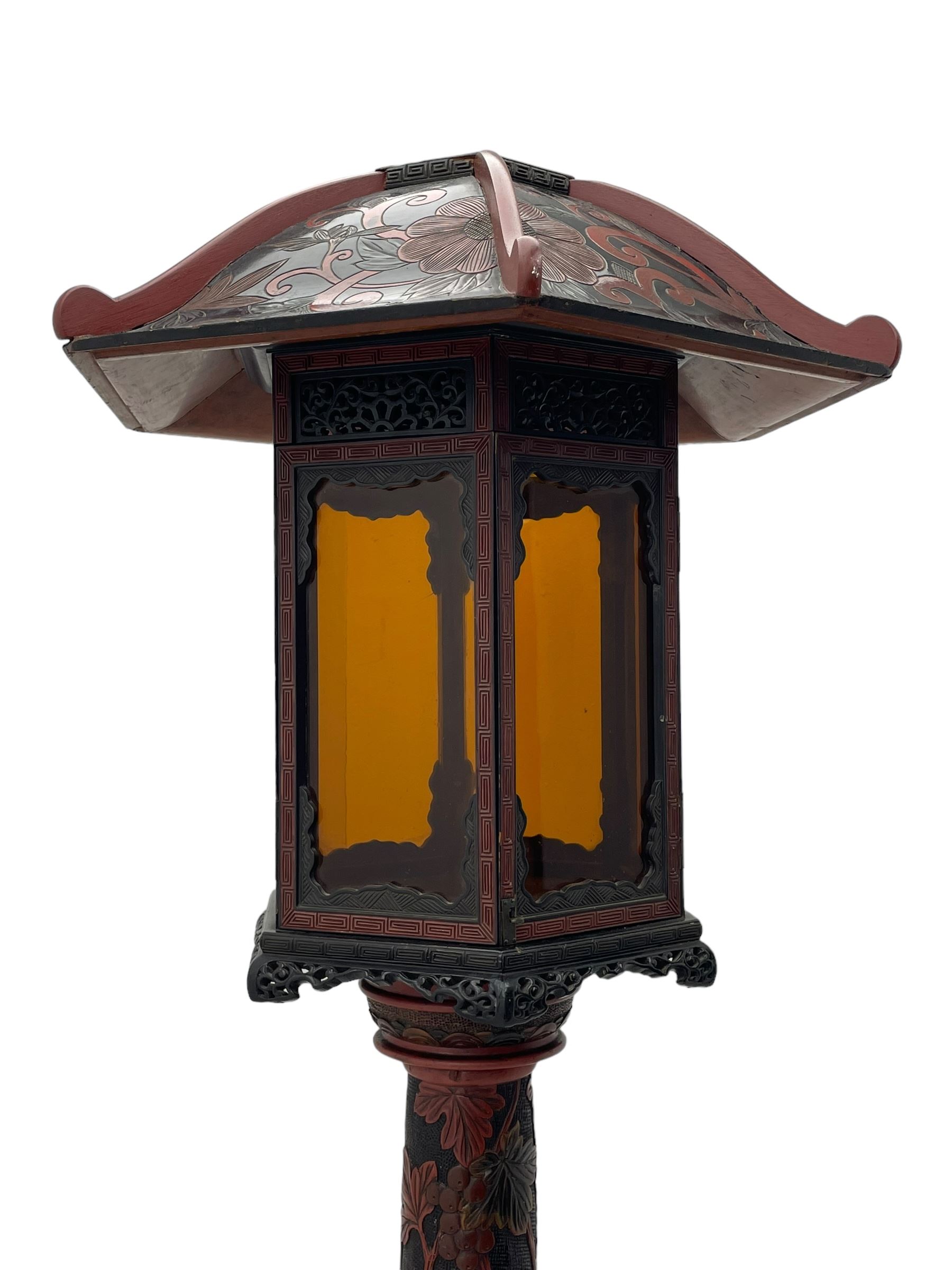 20th century Chinese black and red lacquered hardwood lantern stand, hexagonal shaped canopy top carved with flower heads and scrolls, the lantern fitted with amber tinted glass panes, pierced and carved upper panels, enclosed by two hinged doors, turned stem carved in relief with trailing vine leaves with fruits, stepped hexagonal base with carved decoration  