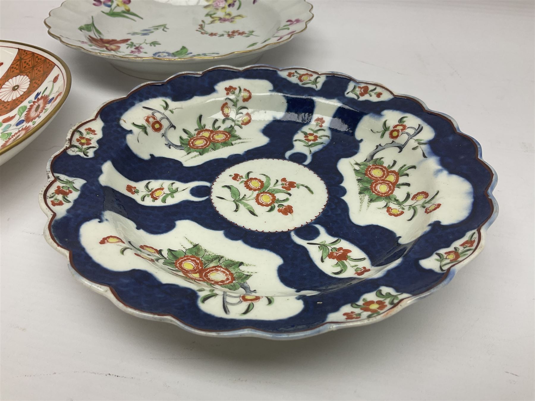 Three 18th century Worcester desert dishes, the first example decorated in the Scarlet Japan pattern, circa 1790, painted with alternating panels of Kakiemon style prunus blossom and chrysanthemum, and orange ground panels with gilt fret detail, D19cm, the second example, circa 1770, decorated with Kakiemon style chrysanthemums and foliage within shaped reserves upon a blue scale ground, with fretted square pseudo mark beneath, D19cm, and the third, circa 1770, painted with polychrome floral sprays and sprigs, D20cm