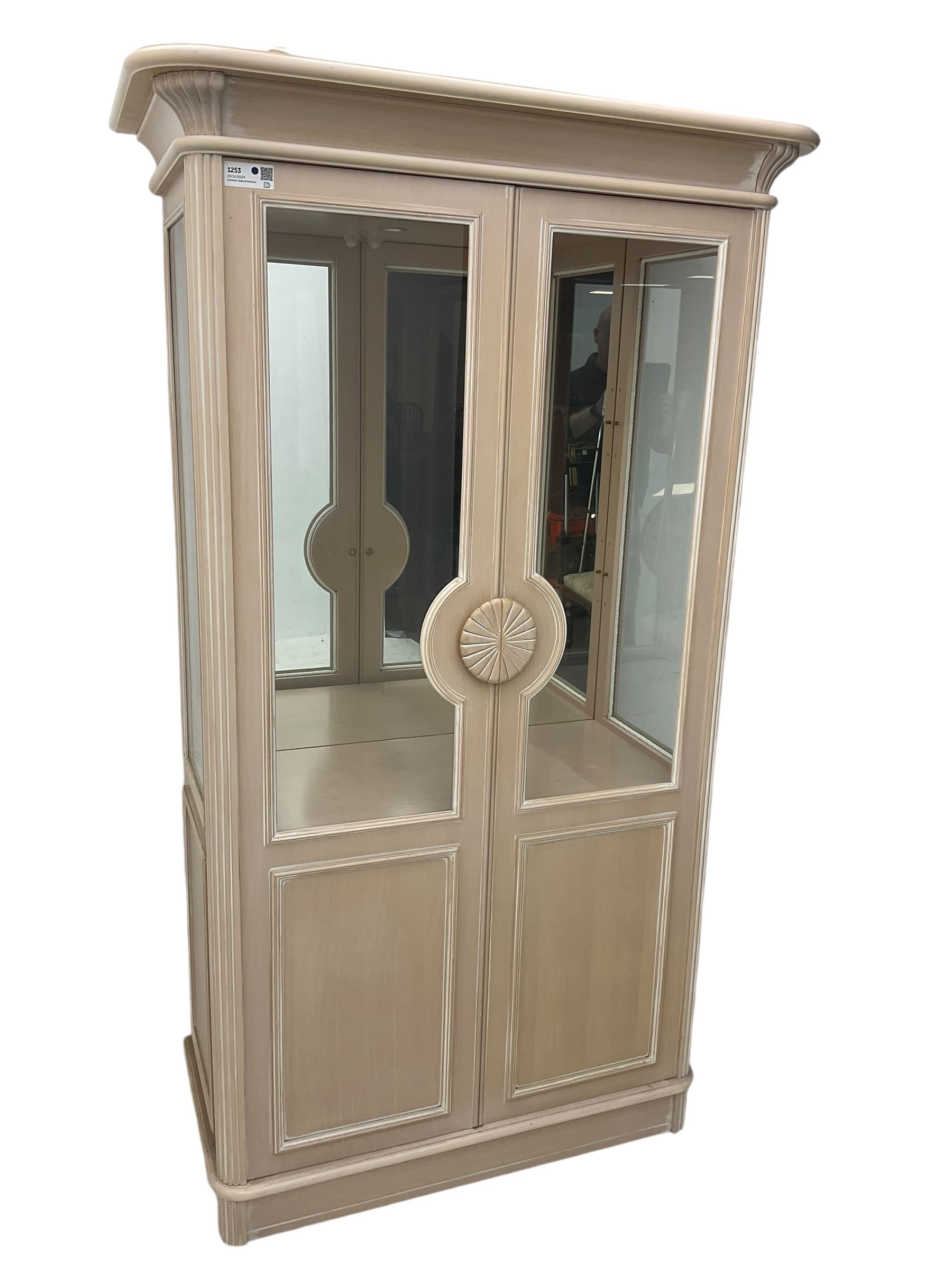 Contemporary display cabinet, moulded cornice above glazed panelled doors with central carved sunburst handles, mirrored back interior with lighting, lower enclosed storage compartment, raised on plinth base