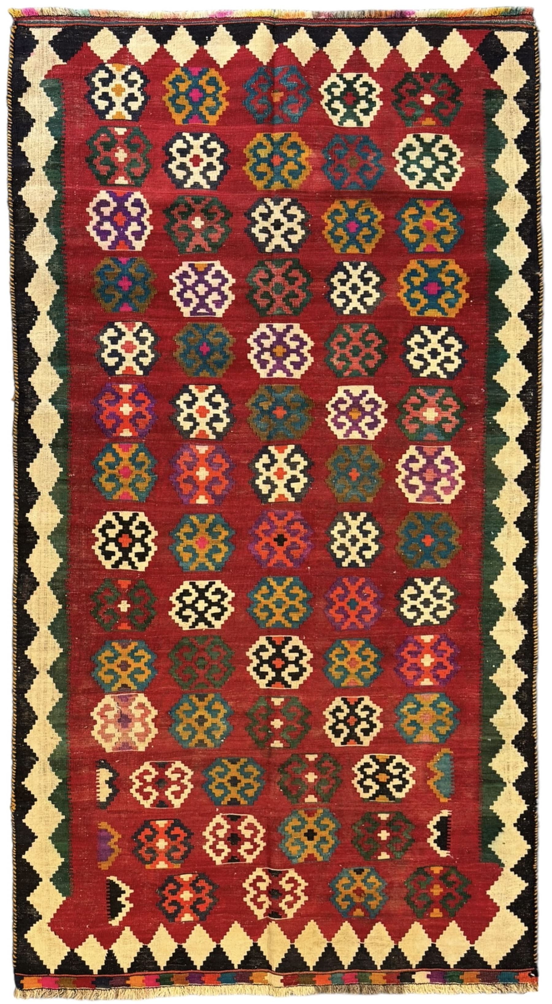 Southwest Persian Qashqai Kilim red ground rug, the central field adorned with rows of multicolored hexagonal medallions with geometric patterns, the border featuring alternating black and white stepped diamonds, narrow band to one end decorated with multicoloured geometric figures