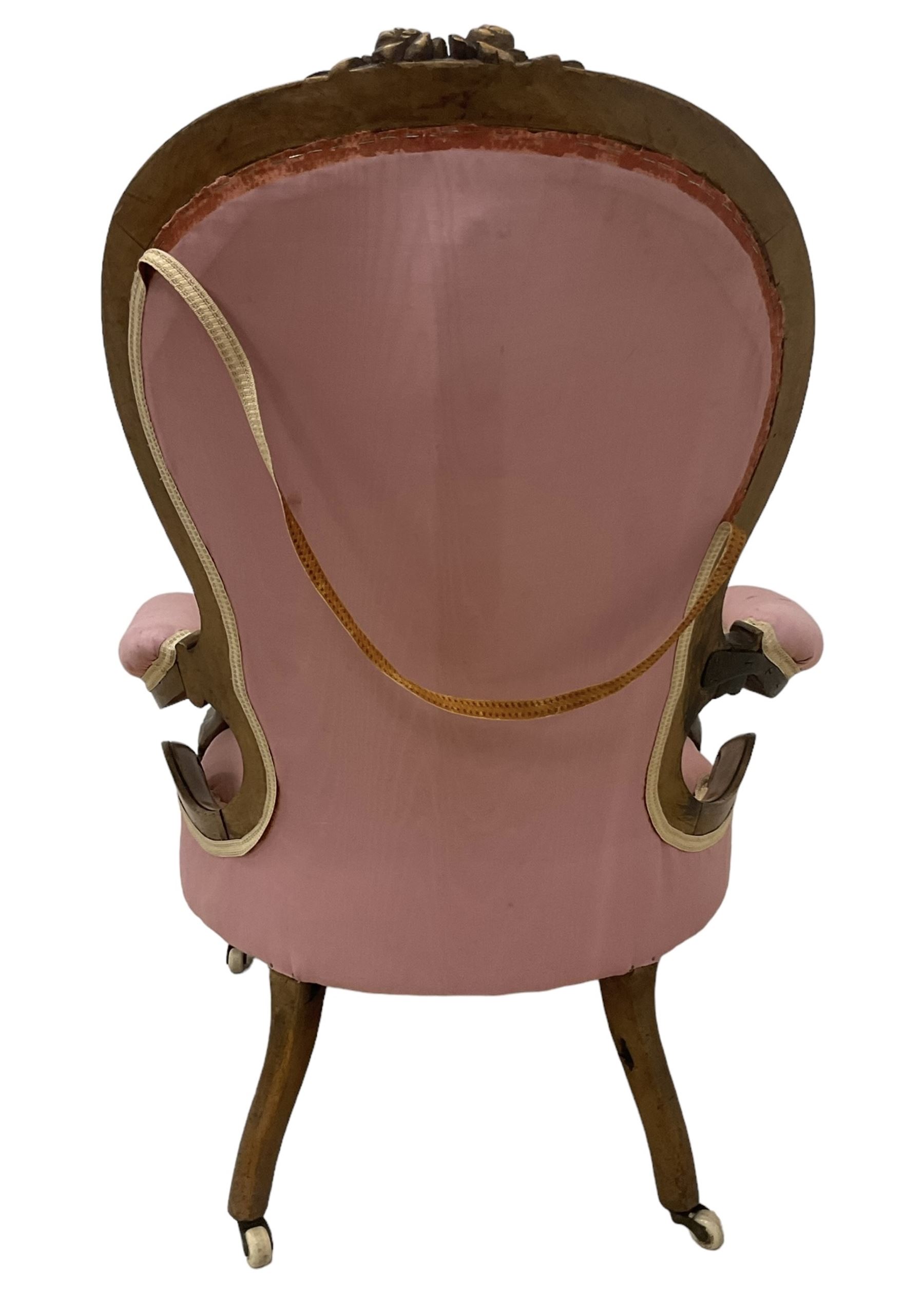 Victorian mahogany framed open armchair, spoon-back and sprung seat upholstered in pink fabric, scrolled arm terminals over serpentine fronted seat decorated with moulded carved floral motifs, cabriole supports with ceramic castors