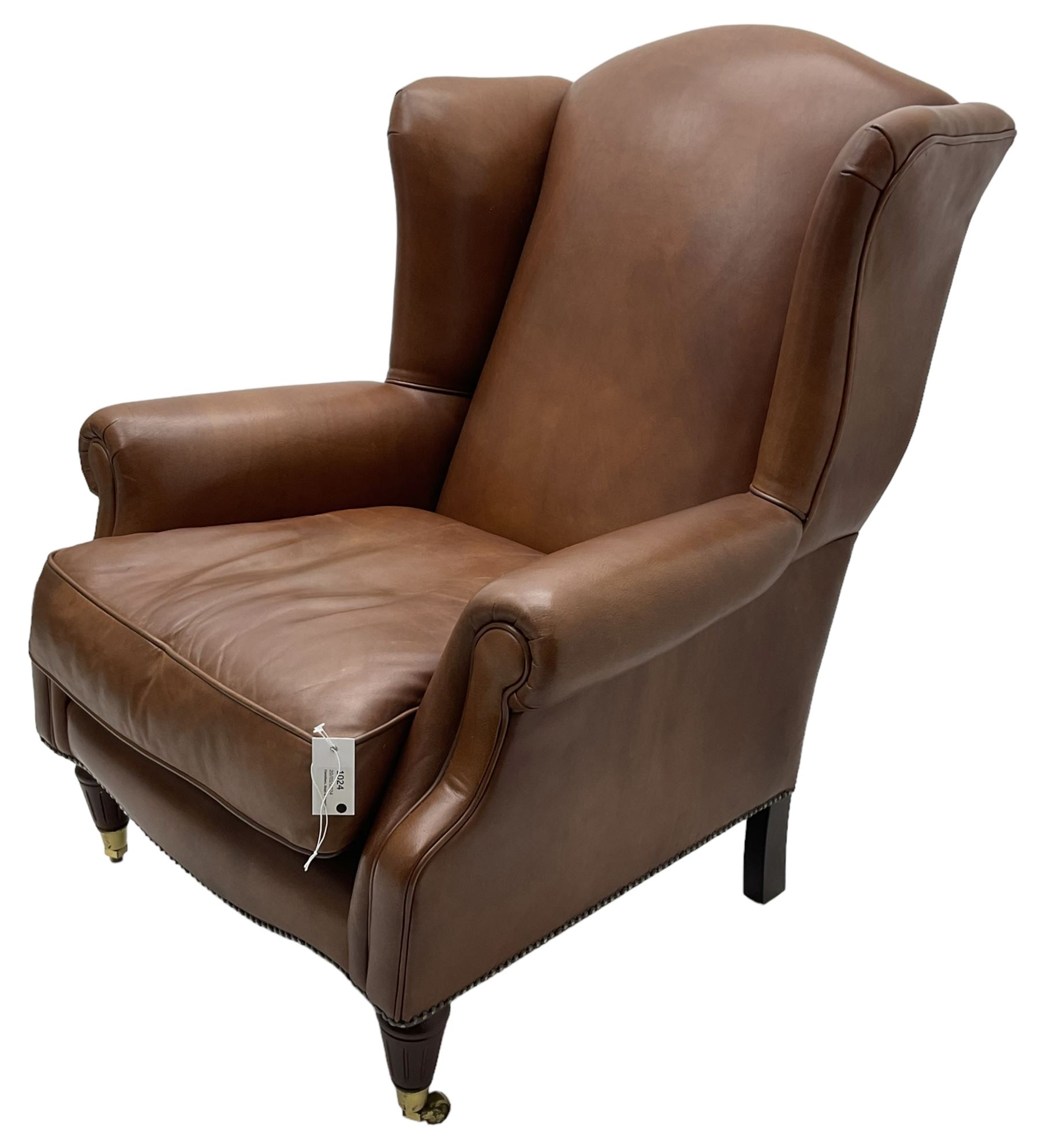 Georgian design wingback armchair, upholstered in brown leather with stud band and piping, turned and fluted front feet on brass cups and castors