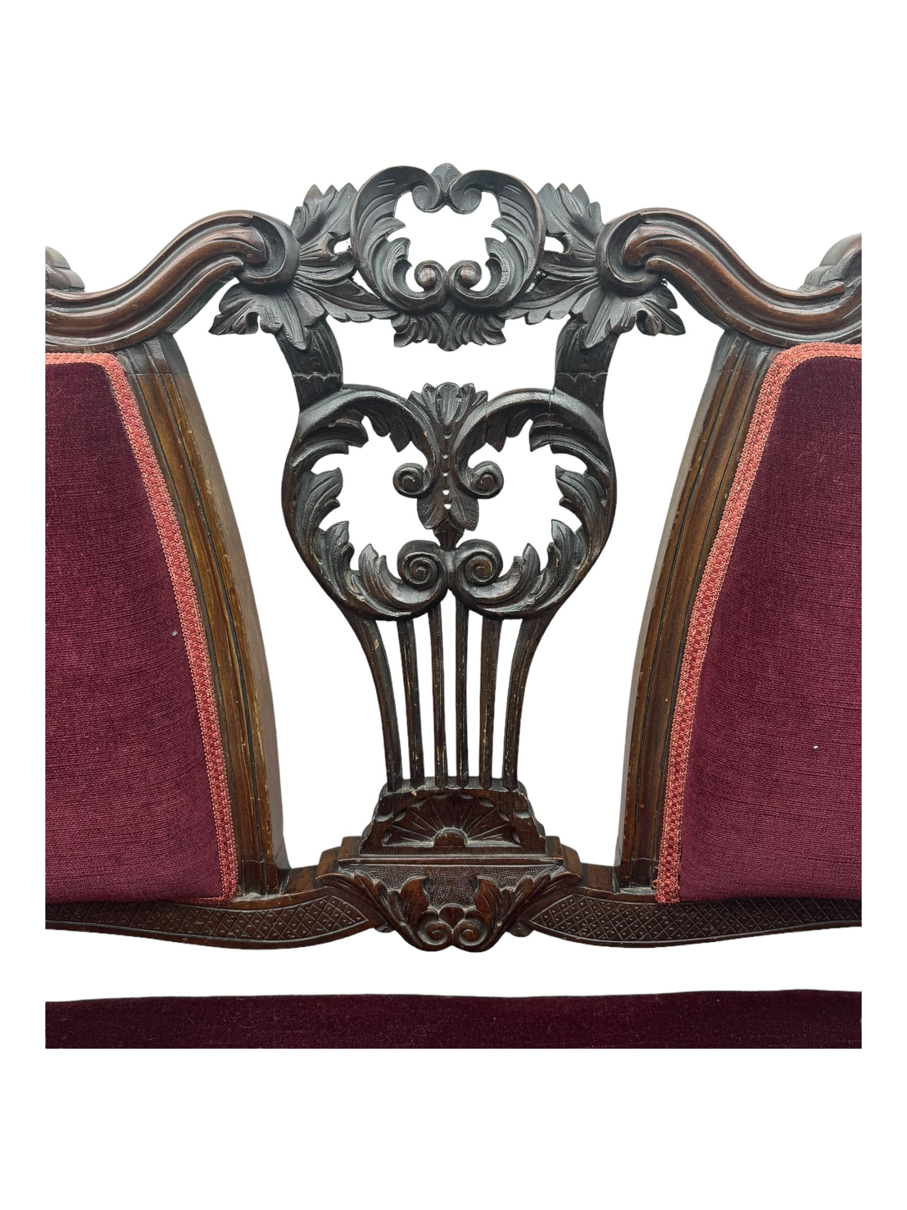 Late Victorian dark oak framed sofa, the backrests, seat and padded arms upholstered in deep red fabric, carved crest rail with scroll and foliate motifs supported by carved uprights, on cabriole supports with leaf carvings terminating in castors