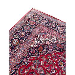 Large Persian Kashan crimson ground carpet, central floral medallion surrounded by swirling leafy branches and palmettes, enclose by floral pattern spandrels, the indigo border with overall scrolling design decorated with palmettes, within guard stripes 